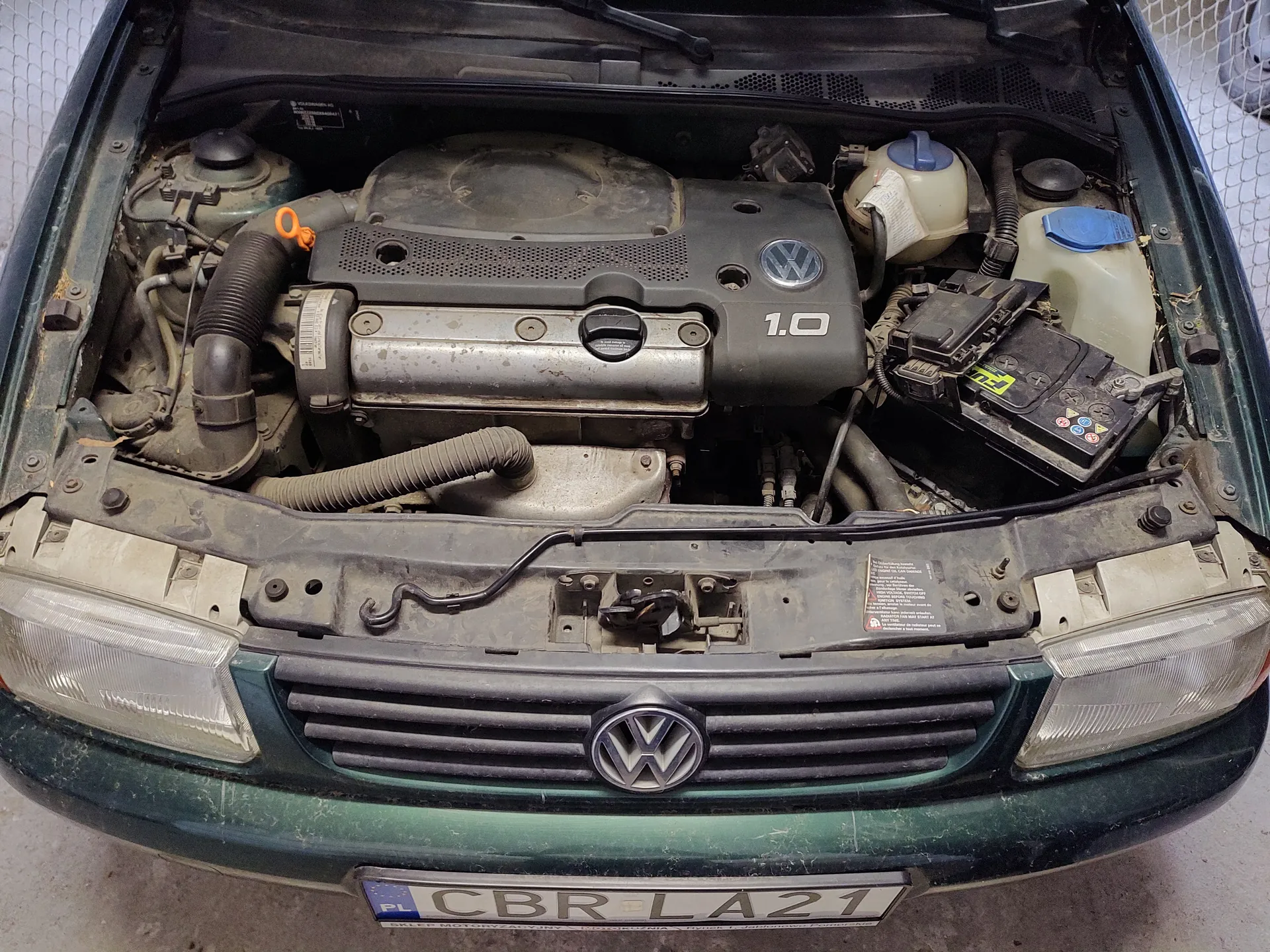 Samochód marki Volkswagen z otwartą maską, ukazujący silnik oznaczony jako 1.0. Widoczne są elementy silnika, w tym zbiorniki z niebieskimi korkami, akumulator, przewody i logo VW na masce. Na dole tablica rejestracyjna z napisem CBR LA21 oraz flagą PL.