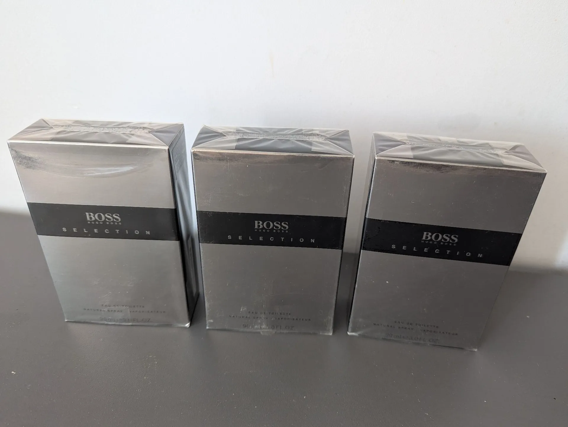 Trzy identyczne, zapakowane w folię pudełka perfum Hugo Boss Selection. Pudełka są w kolorze ciemnoszarym z czarnym pasem w połowie wysokości, na którym znajduje się biały napis 'BOSS HUGO BOSS' oraz poniżej słowo 'SELECTION'. Na dole pudełek widnieje mniejszy tekst: 'EAU DE TOILETTE NATURAL SPRAY VAPORISATEUR 90ml e 3.0 FL.OZ.'. Pudełka stoją obok siebie na szarej powierzchni na tle białej ściany.