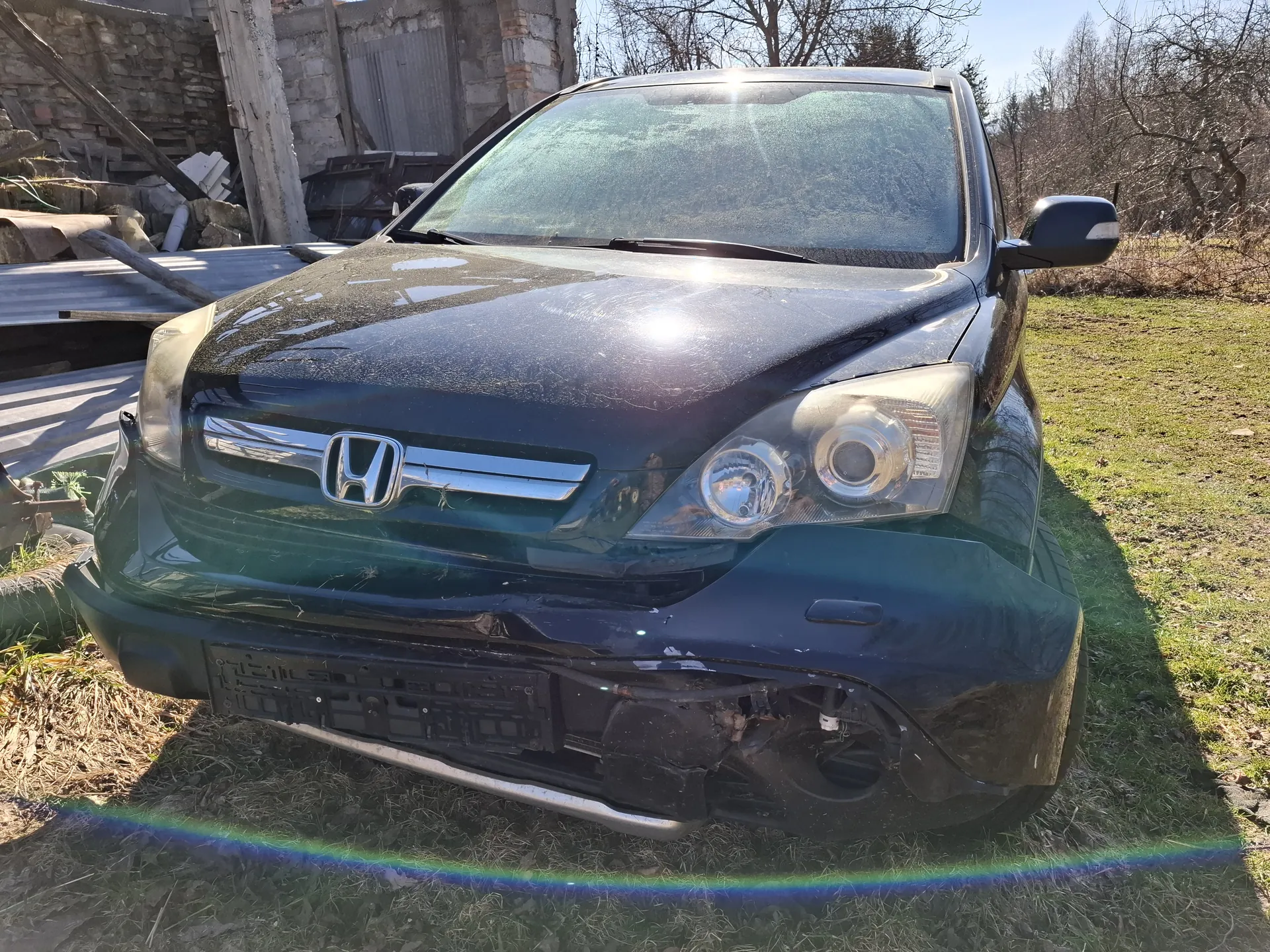 Czarny samochód marki Honda z uszkodzonym przodem, w tym zdeformowanym zderzakiem i uszkodzonymi reflektorami. Samochód stoi na trawie, na tle budynku w stanie ruin oraz stosów materiałów budowlanych. Słoneczny dzień, widoczne odbicia słońca na masce.