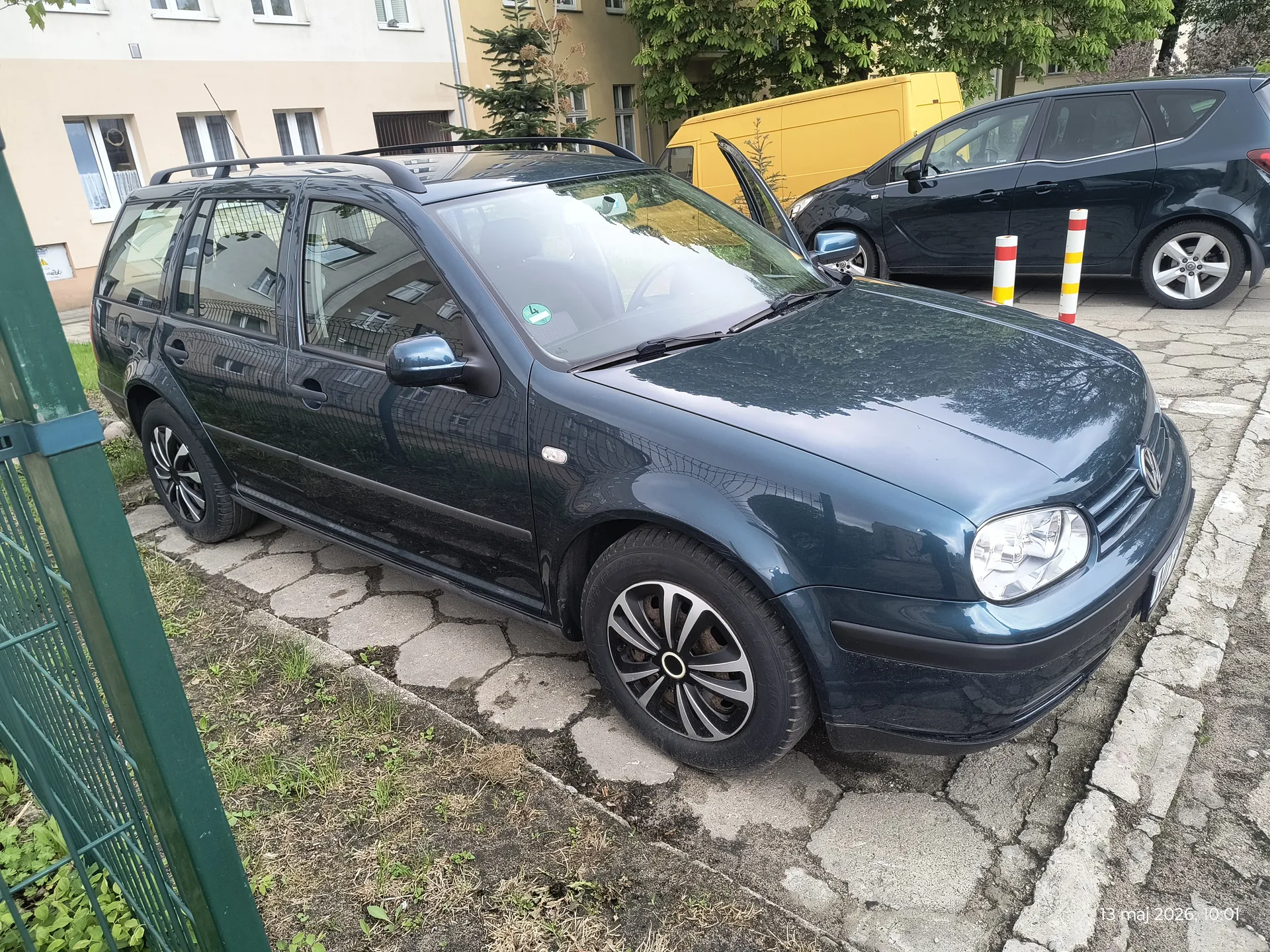 Ciemnozielony samochód typu kombi zaparkowany na brukowanej powierzchni. Po lewej stronie zielone ogrodzenie, w tle czarny samochód z otwartą przednią drzwią, żółty van oraz budynki mieszkalne z oknami. Na pierwszym planie widoczne są kolorowe słupki (czerwono-żółte) oraz drzewa. W prawym dolnym rogu znacznik daty: 13 maj 2026, 10:01. Samochód posiada dachowy bagażnik i naklejkę na przedniej szybie.