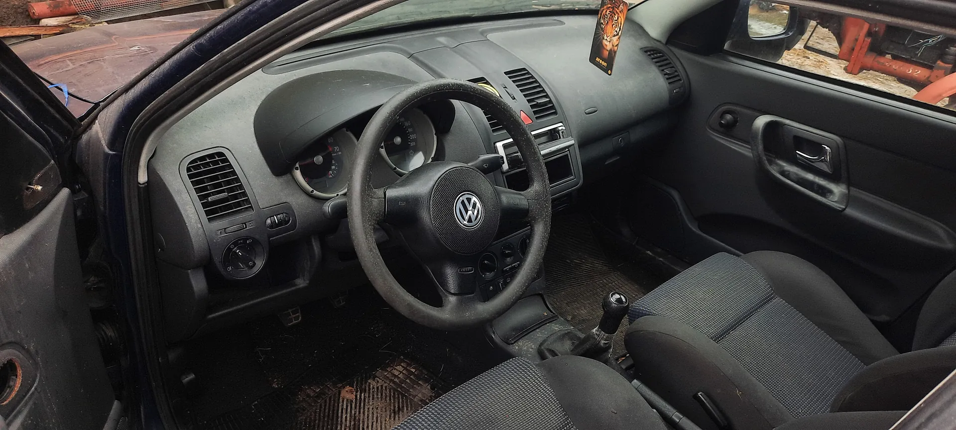 Wnętrze samochodu Volkswagen z czarną kierownicą z logo VW, pokrytą kurzem deską rozdzielczą, licznikami, przełącznikami klimatyzacji, dźwignią biegów, siedzeniami tkaninowymi, zanieczyszczonymi podłogami i wiszącym odświeżaczem z obrazem tygrysa. Drzwi kierowcy otwarte.
