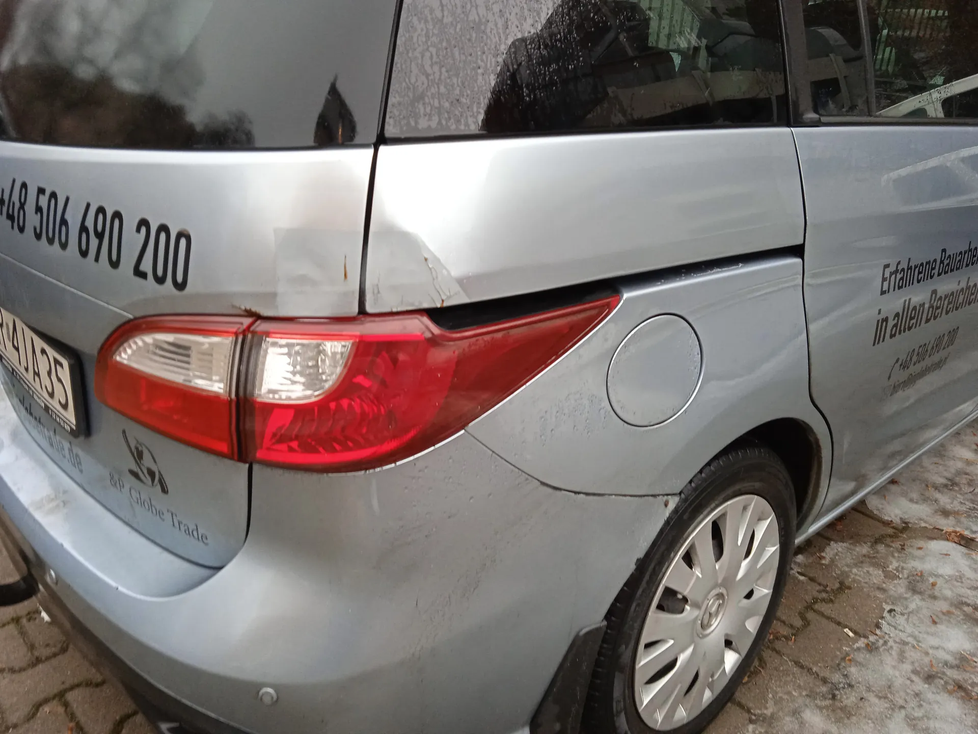 Zbliżenie na tylną i boczną część srebrnego furgonu. Na tylnej szybie widoczny czarny numer telefonu +48 506 690 200. Na tylnej klapie znajduje się biały numer rejestracyjny R4JA35, logo oraz napis B&P Globe Trade. Na boku furgonu umieszczono niemiecki napis Erfahrene Bauarbe in allen Bereichen oraz powtórzony numer telefonu +48 506 690 200. Furgon stoi na brukowanej powierzchni z resztkami śniegu lub lodu.