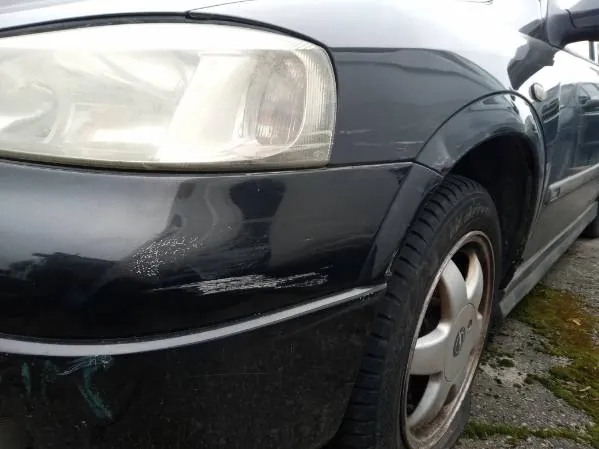 Czarny samochód widziany z przodu po lewej stronie. Widoczny jest reflektor, przedni zderzak z widoczną zadrapiną, koło z srebrną felgą oraz fragment boku pojazdu. Samochód stoi na nierównym podłożu z drobnym żwirem i trawą. Na karoserii widoczne są ślady brudu lub kropli wody.