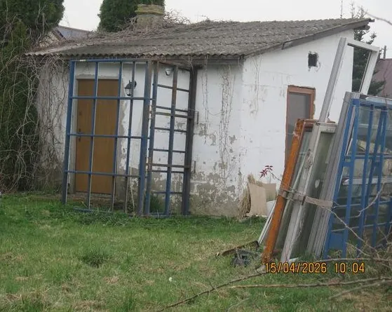Mały, biały budynek gospodarczy z uszkodzoną elewacją i dwuspadowym dachem z płyt falistych. Przed budynkiem znajdują się niebieskie metalowe ramy okienne lub drzwiowe, z których jedna jest oparta o ścianę, a pozostałe leżą lub stoją obok siebie na trawiastym terenie. W prawym dolnym rogu widnieje pomarańczowy napis z datą i godziną: 15/04/2026 10:04.