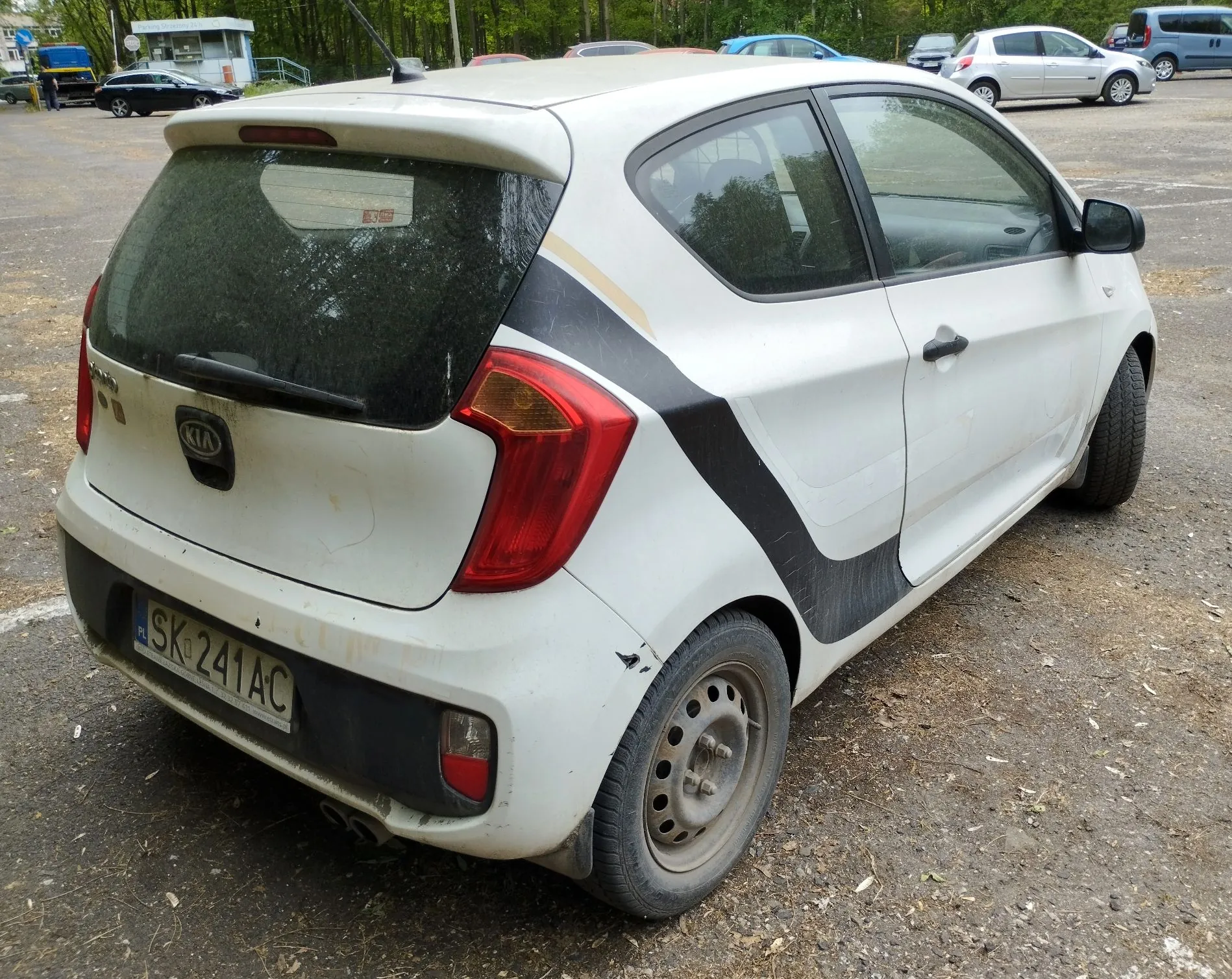 Biały samochód marki Kia w nadwoziu hatchback, zaparkowany na nieutwardzonym placu. Pojazd ma czarne pasy dekoracyjne na tylnym błotniku i drzwiach oraz widoczne zabrudzenia na karoserii. Tablica rejestracyjna to SK 241AC. W tle widoczne są inne zaparkowane samochody oraz budynek.