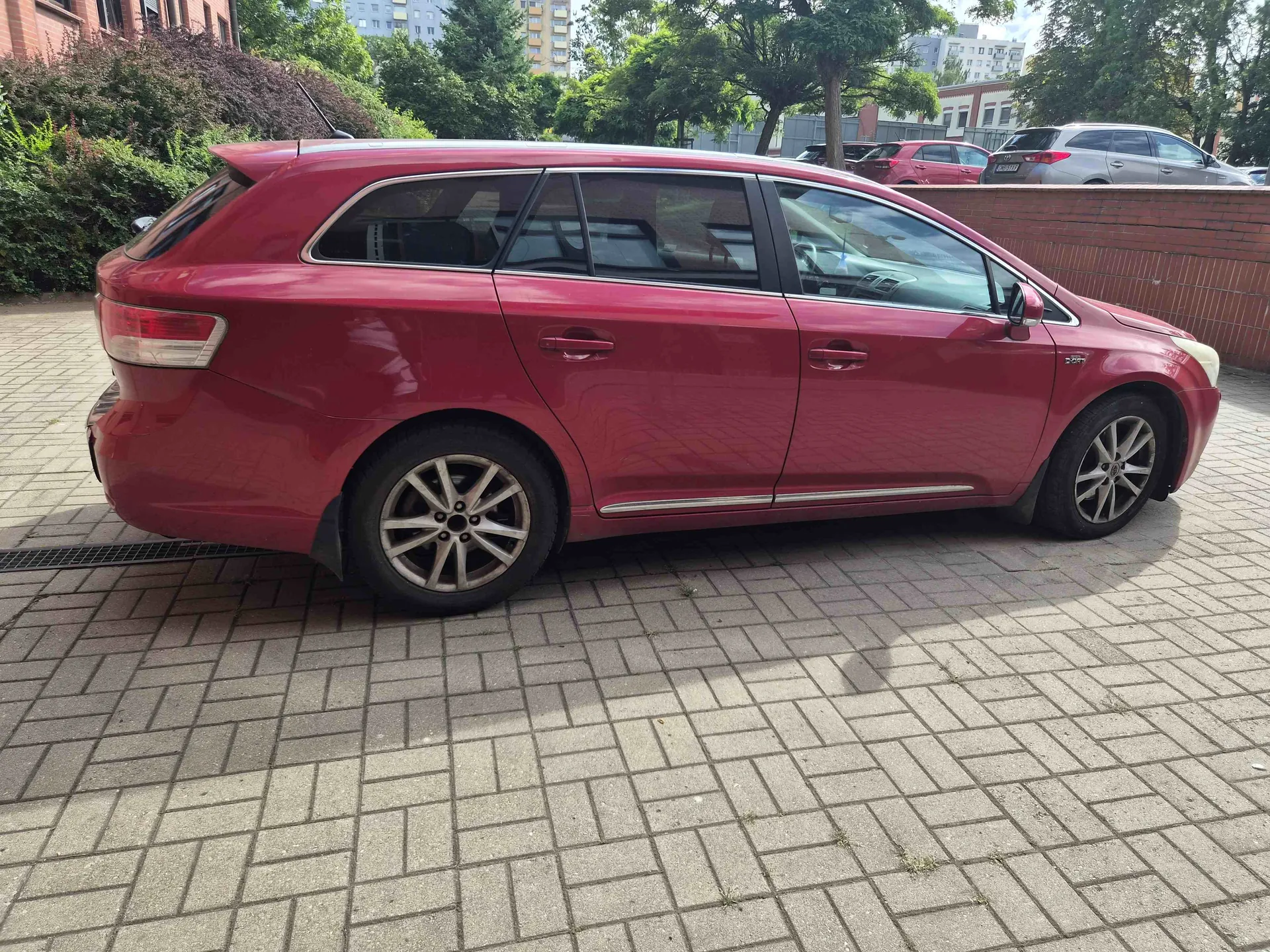 Profil czerwonego samochodu typu kombi zaparkowanego na kostce brukowej. W tle widoczne są zielone krzewy, drzewa oraz fragmenty budynków mieszkalnych i inne zaparkowane samochody.