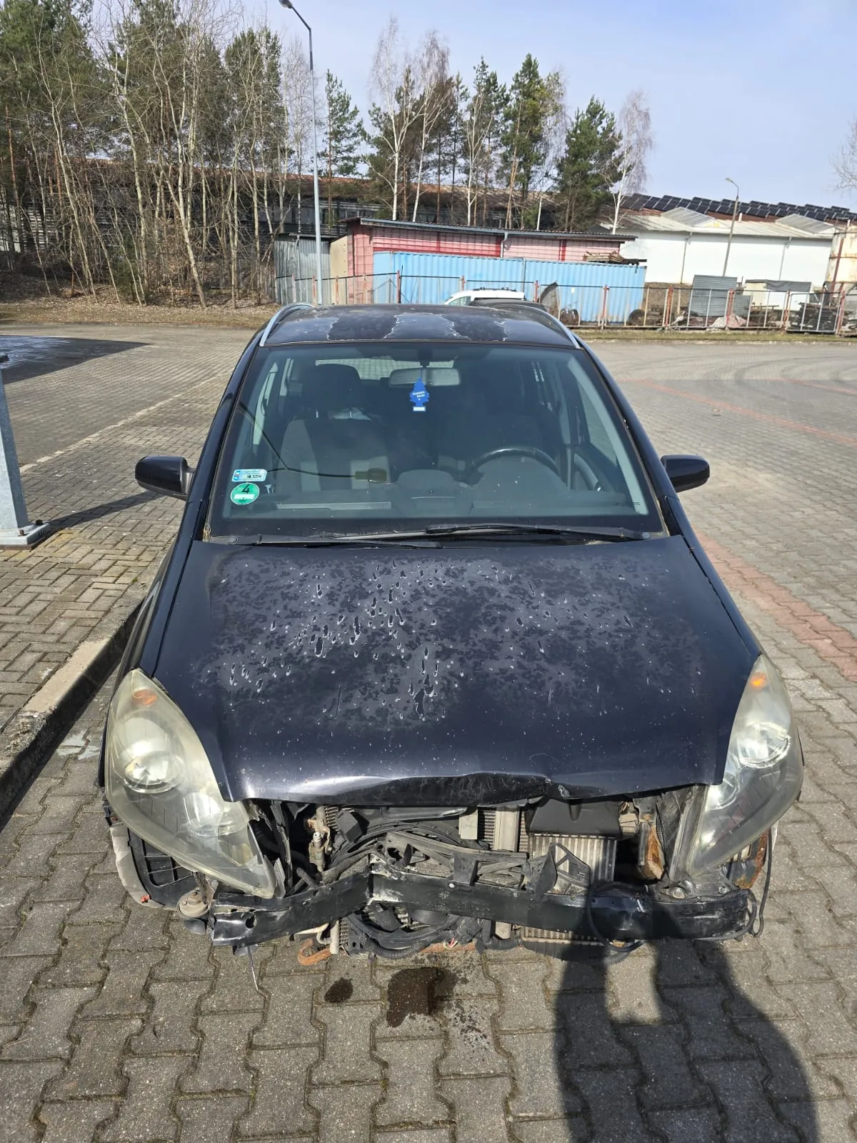 Czarny samochód osobowy z uszkodzonym przodem, zdemontowanym zderzakiem i odsłoniętymi częściami silnika. Na masce widoczne krople wody. Samochód zaparkowany na brukowanej powierzchni. W tle drzewa, budynki przemysłowe, niebieski kontener oraz słoneczna pogoda. Na przedniej szybie naklejki, w tym zielona z cyfrą 4.