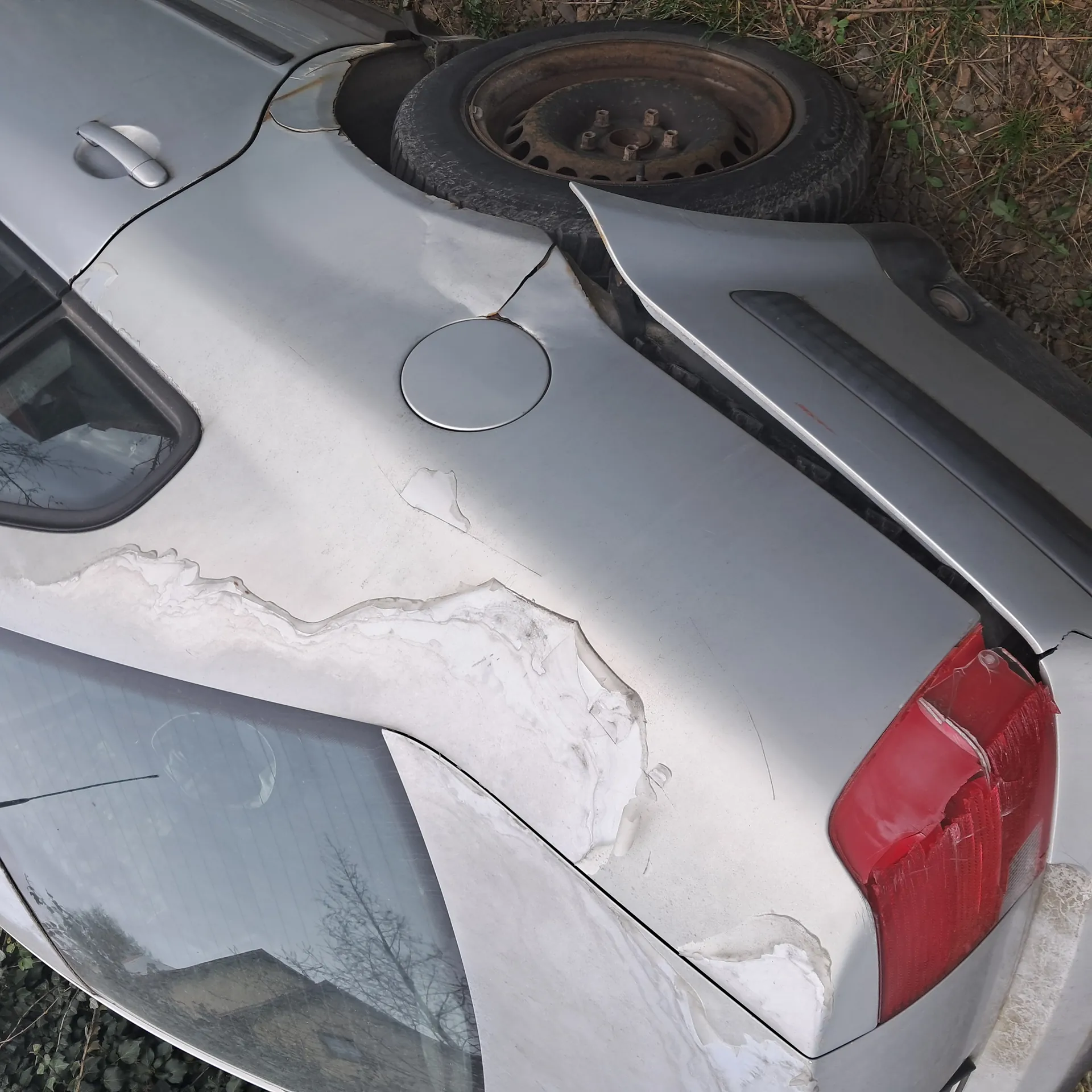 Zdjęcie przedstawia uszkodzony srebrny samochód widziany z góry. Widoczne są poważne deformacje nadwozia w tylnej części, w tym pęknięcia i zarysowane miejsca z odsłoniętą białą podkładką. Czerwone światło tylnego reflektora jest pęknięte. Obok samochodu leży stary, rdzewiejący felga z oponą. Tło stanowi ziemia z trawą i suchymi liśćmi.