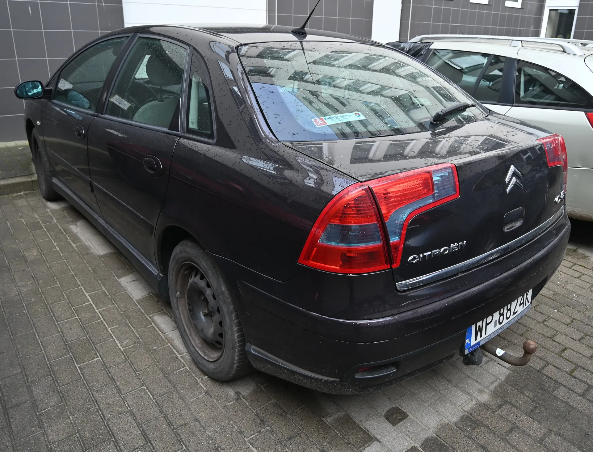 Czarny samochód marki Citroën C5 zaparkowany na brukowanej powierzchni. Widok z tyłu, z widocznymi czerwonymi światłami tylnymi, tablicą rejestracyjną WP 8824K z niebieskim pasem PL po lewej stronie, logo Citroën i napisem C5 na bagażniku. Na tylnej szybie naklejki, a na dachu antena. W tle budynek z szarymi płytkami i srebrny samochód. Powierzchnia brukowa i wilgotna karoseria wskazują na niedawny deszcz.