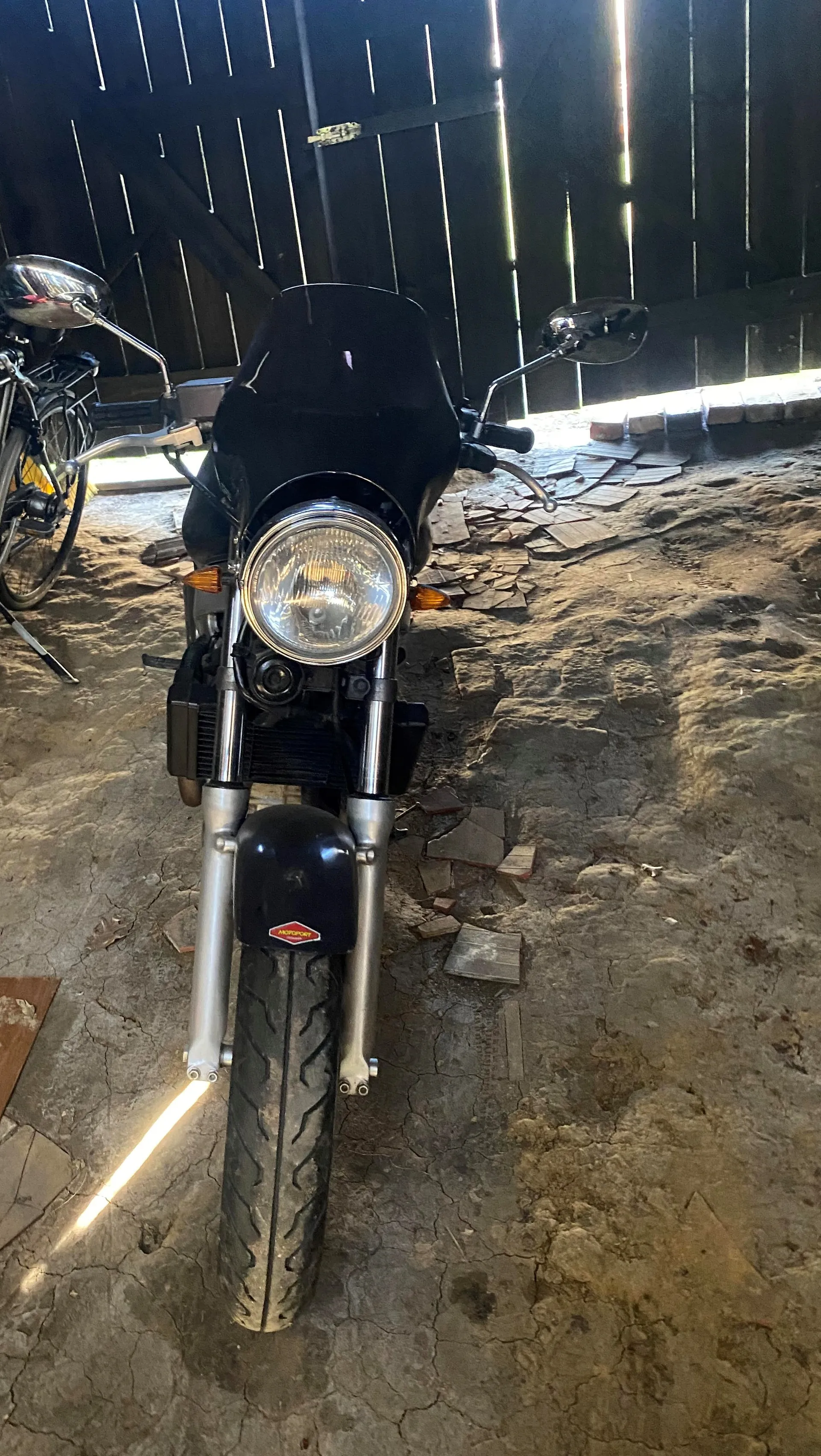 Przód czarnego motocykla z dużym okrągłym reflektorem, lusterkami bocznymi i czerwonym emblematem na przednim błotniku. Motocykl stoi na betonowej podłodze z rozsypanymi kawałkami płytek. W tle ciemne drewniane ściany budynku z pionowymi deskami. Po lewej stronie częściowo widoczny rower z metalowym lusterkiem.