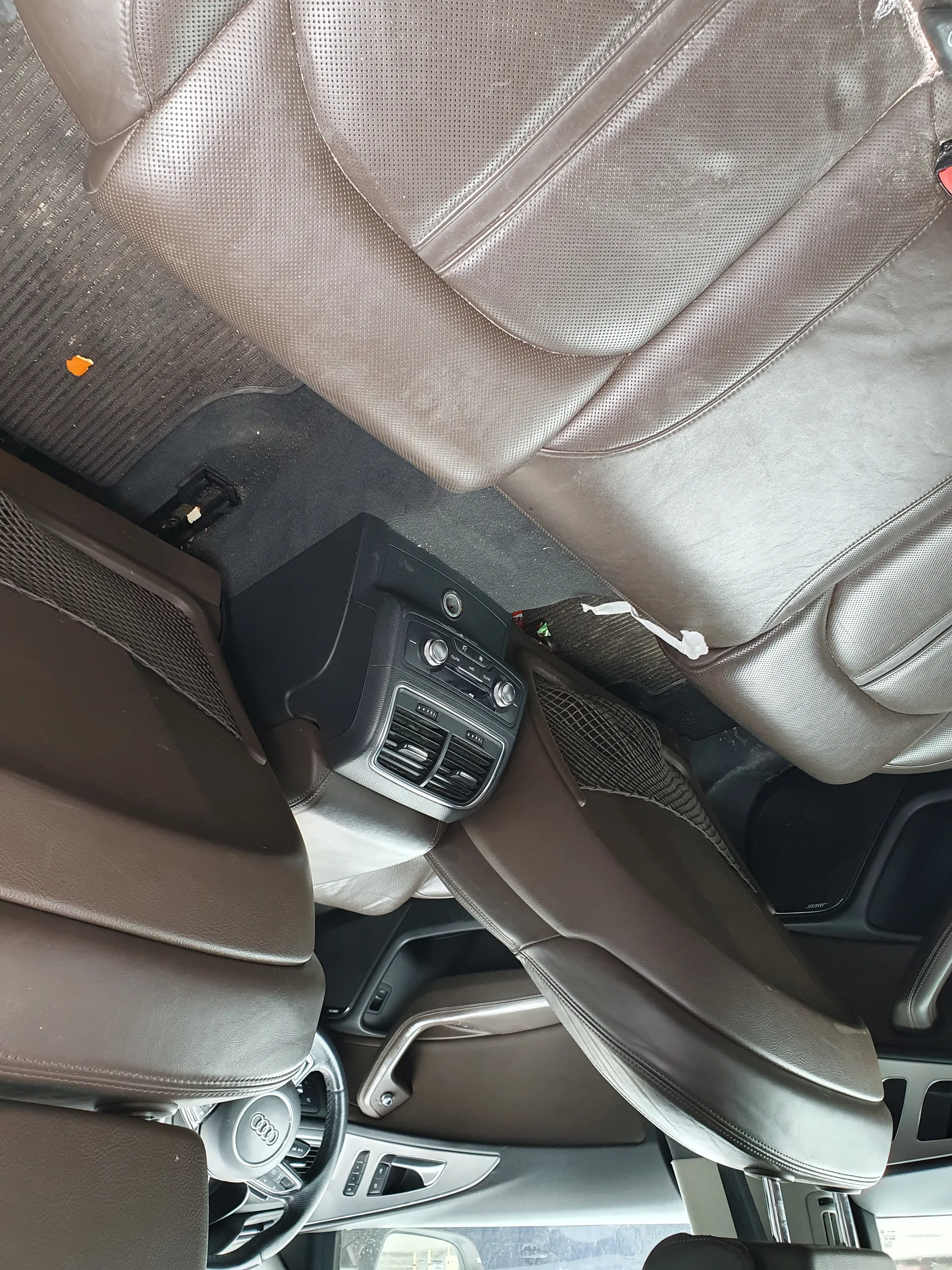 Wnętrze samochodu marki Audi z brązowymi skórzany siedzeniami. Widoczna kierownica z logo Audi, deska rozdzielcza, centralna konsola z wentylacją i przyciskami sterującymi. Na podłodze dywanik z małym pomarańczowym przedmiotem oraz śladami brudu. Fragmenty siedzeń i drzwi z elementami wykończeniowymi w kolorze brązowym.
