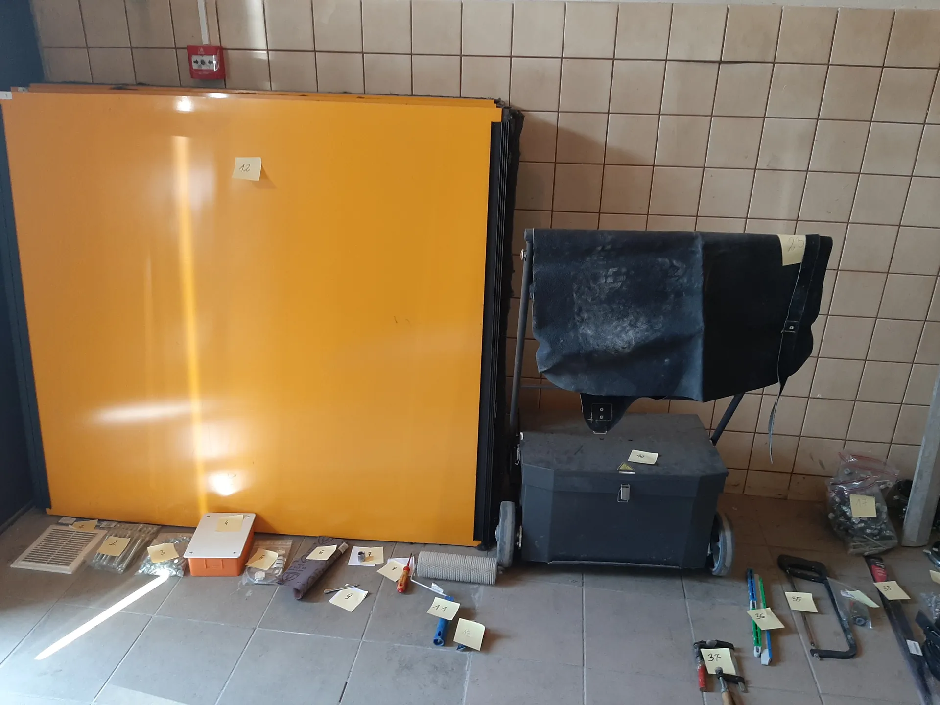 Pomieszczenie z beżową płytkową ścianą i szarym podłogowym płytkami. Po lewej stronie duży pomarańczowy panel oparty o ścianę z naklejką oznaczoną numerem 12. Obok czarny wózek z kółkami, na którym znajduje się czarna skrzynia i ciemny materiał. Na podłodze rozłożone narzędzia i przedmioty z białymi etykietami oznaczonymi numerami: 3, 4, 5, 6, 7, 8, 9, 11, 13, 23, 35, 36, 37. W lewym górnym rogu czerwony pożarniczy przycisk. W tle widoczne opakowania z małymi przedmiotami.