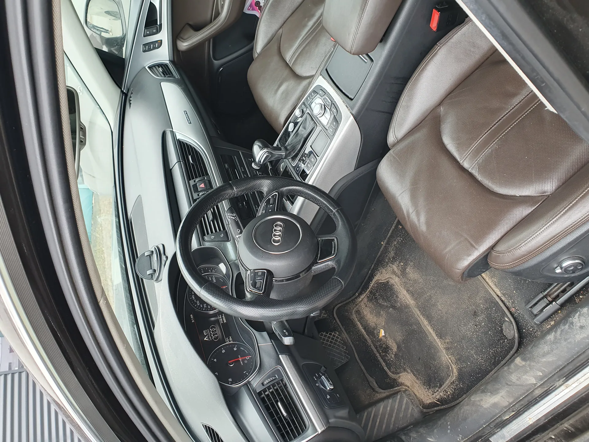 Wnętrze samochodu marki Audi z brązowymi skórzany fotelami, kierownicą z logo Audi, deską rozdzielczą z licznikami, dźwignią zmiany biegów oraz zabrudzonymi dywanikami. Drzwi pasażera są otwarte, przez okno widać fragment budynku.