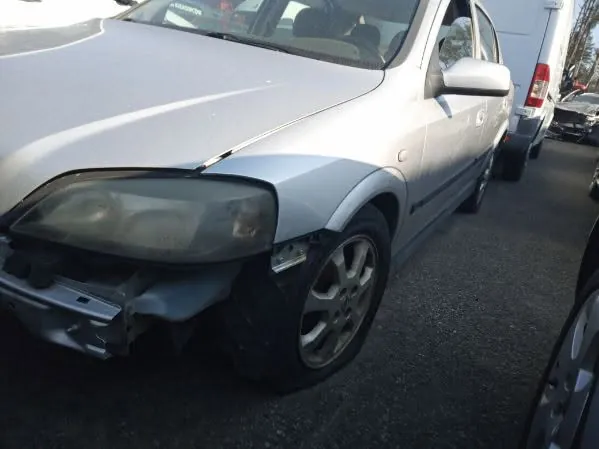 Srebrny samochód osobowy z uszkodzonym przodem. Widoczne pęknięte reflektory, zdeformowany zderzak i uszkodzona maska. Samochód zaparkowany na asfaltowej powierzchni, w tle inne pojazdy, w tym biały van i czarny samochód. Brak czytelnego tekstu na obrazie.