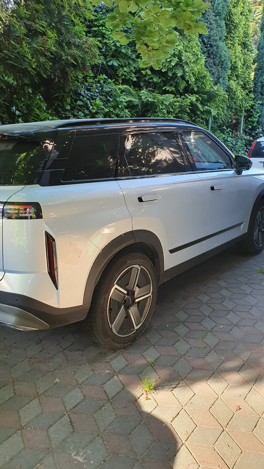 Boczny widok białego samochodu typu SUV zaparkowanego na kostce brukowej. Pojazd ma czarny dach, przyciemniane szyby i charakterystyczne pięcioramienne felgi aluminiowe. W tle widoczne są gęste, zielone krzewy i drzewa.