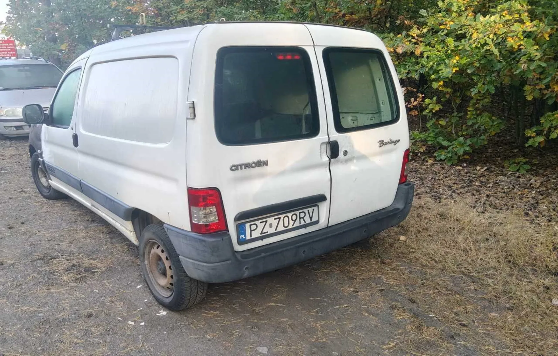 Biały samochód dostawczy marki Citroën Berlingo zaparkowany na szutrowym podłożu na tle zielonych drzew. Widoczny jest tył i lewy bok pojazdu. Samochód posiada polską tablicę rejestracyjną o numerze PZ 709RV. Na klapie bagażnika widnieją napisy CITROËN oraz Berlingo.
