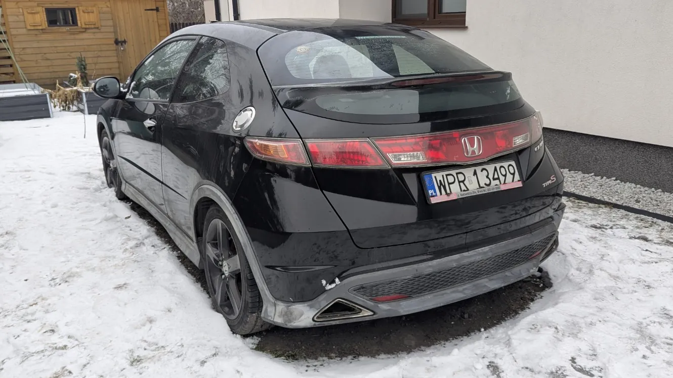 Czarny samochód marki Honda, widok z tyłu, zaparkowany na śnieżnej powierzchni. Widoczna tablica rejestracyjna z polskim kodem PL i numerem WPR 13499. W tle budynek z drewnianymi drzwiami i oknami, a także śnieg pokrywający ziemię. Samochód posiada czerwone światła tylnego oraz logo Hondy na bagażniku.