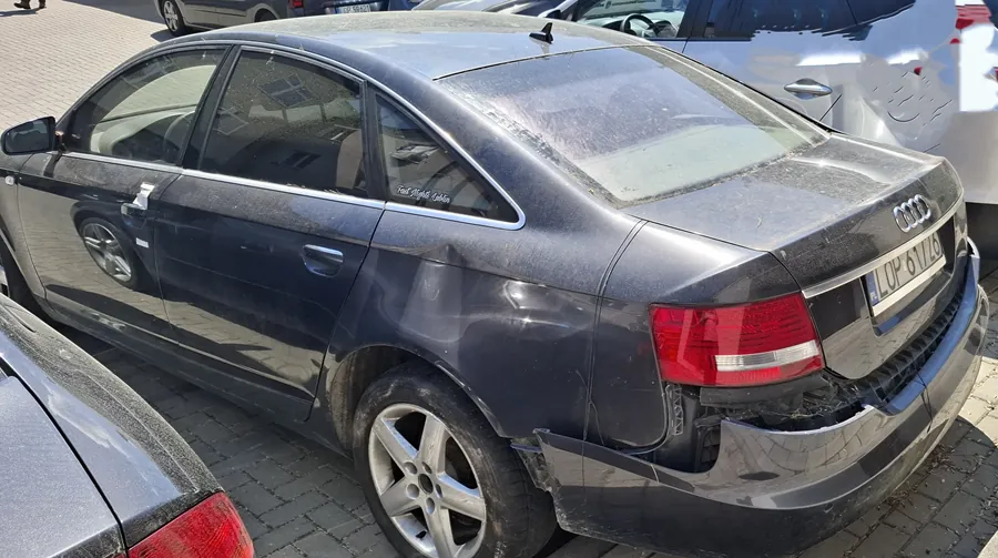 Ciemnoszary samochód Audi sedan zaparkowany na brukowanej powierzchni. Widoczne uszkodzenia na tylnym zderzaku i prawej tylnej części nadwozia. Tablica rejestracyjna z numerem LUP 6V20. W tle częściowo widoczne inne pojazdy, w tym biały samochód. Na tylnym szybie widoczny napis 'Fiat Professional'. Nadwozie pokryte kurzem i zabrudzeniami.