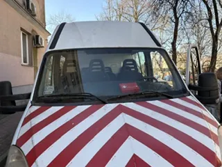 Biały pojazd z czerwonymi i białymi pionowymi pasami w kształcie zygzaka na masce. Widoczne przednie szyby, siedzenia wewnątrz oraz otwarte drzwi po stronie kierowcy. W tle budynki mieszkalne, drzewa bez liści i jasne niebo. Na desce rozdzielczej widoczny czerwony znak.