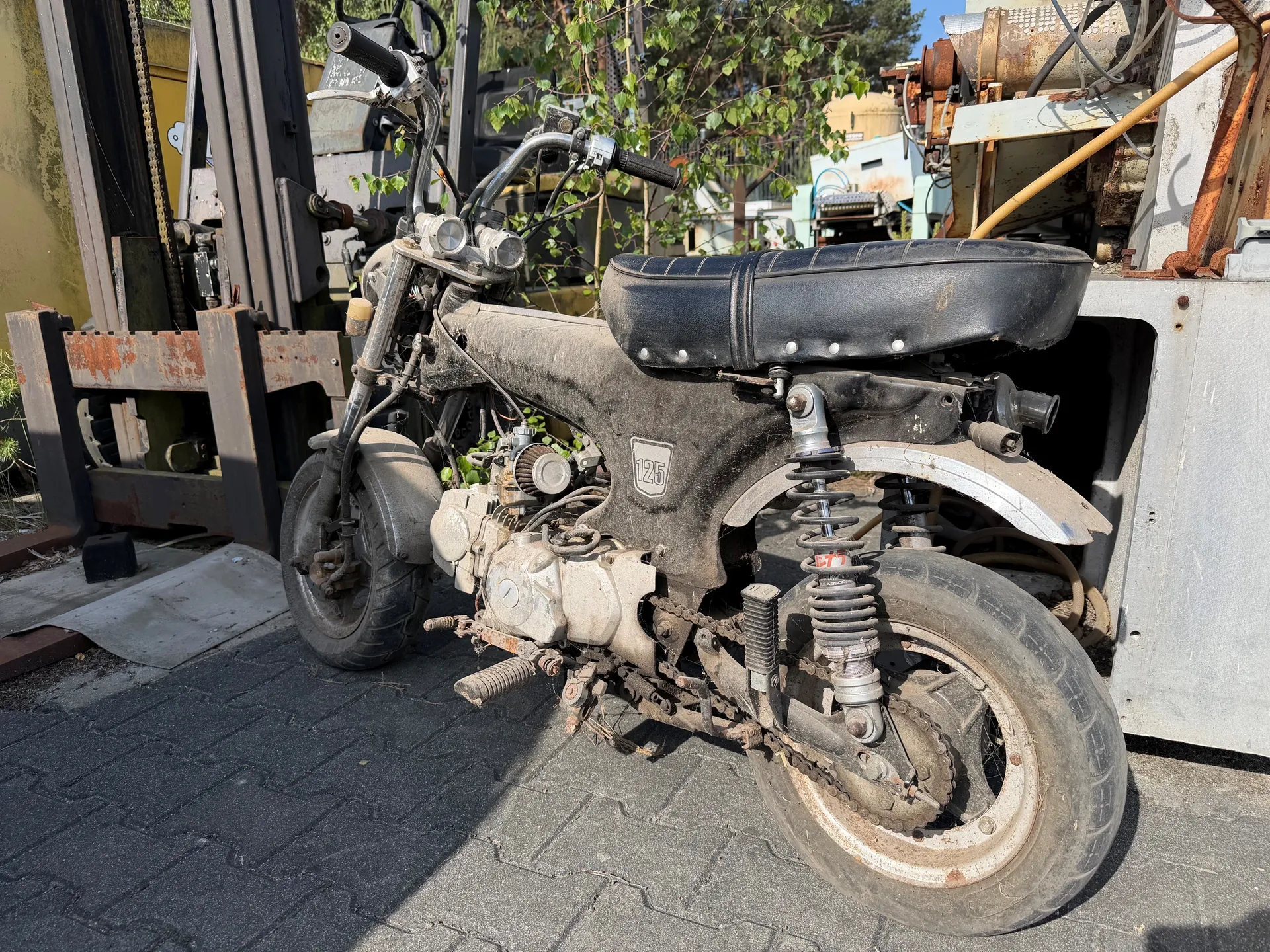 Stary, zardzewiały motocykl typu moped z czarnym, zużytym siedzeniem, pokryty kurzem. Na boku widoczny biały napis "125". Motocykl stoi na brukowanej powierzchni, w tle przemysłowe urządzenia, w tym rostwierdzone maszyny i fragmenty pojazdów. W oddali drzewa i zielone liście. Słoneczny dzień, cienie wskazują na jasne oświetlenie.