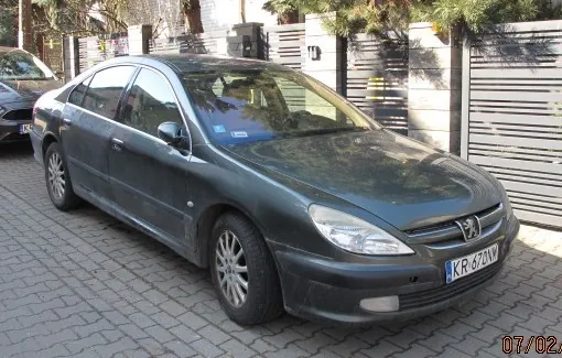 Ciemnozielony samochód osobowy marki Peugeot zaparkowany na brukowanej ulicy. Widoczna tablica rejestracyjna KR 470W. W tle drewniane ogrodzenie z pionowymi listwami, drzewa oraz częściowo widoczny inny samochód po lewej stronie. W prawym dolnym rogu obrazu znajduje się data 07/02.