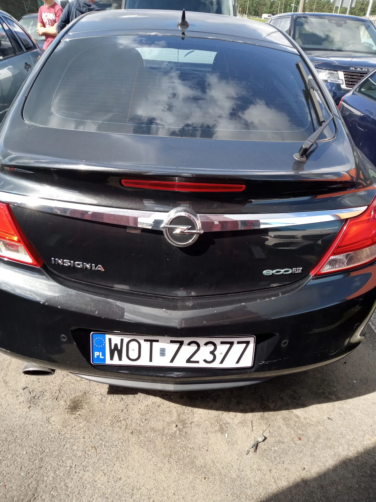 Tylny widok czarnego samochodu marki Opel Insignia. Na klapie bagażnika widoczne jest logo Opela, napis 'INSIGNIA' po lewej stronie oraz 'ecoFLEX' po prawej. Samochód posiada polską tablicę rejestracyjną o numerze WOT 72377. W tylnej szybie odbijają się chmury.