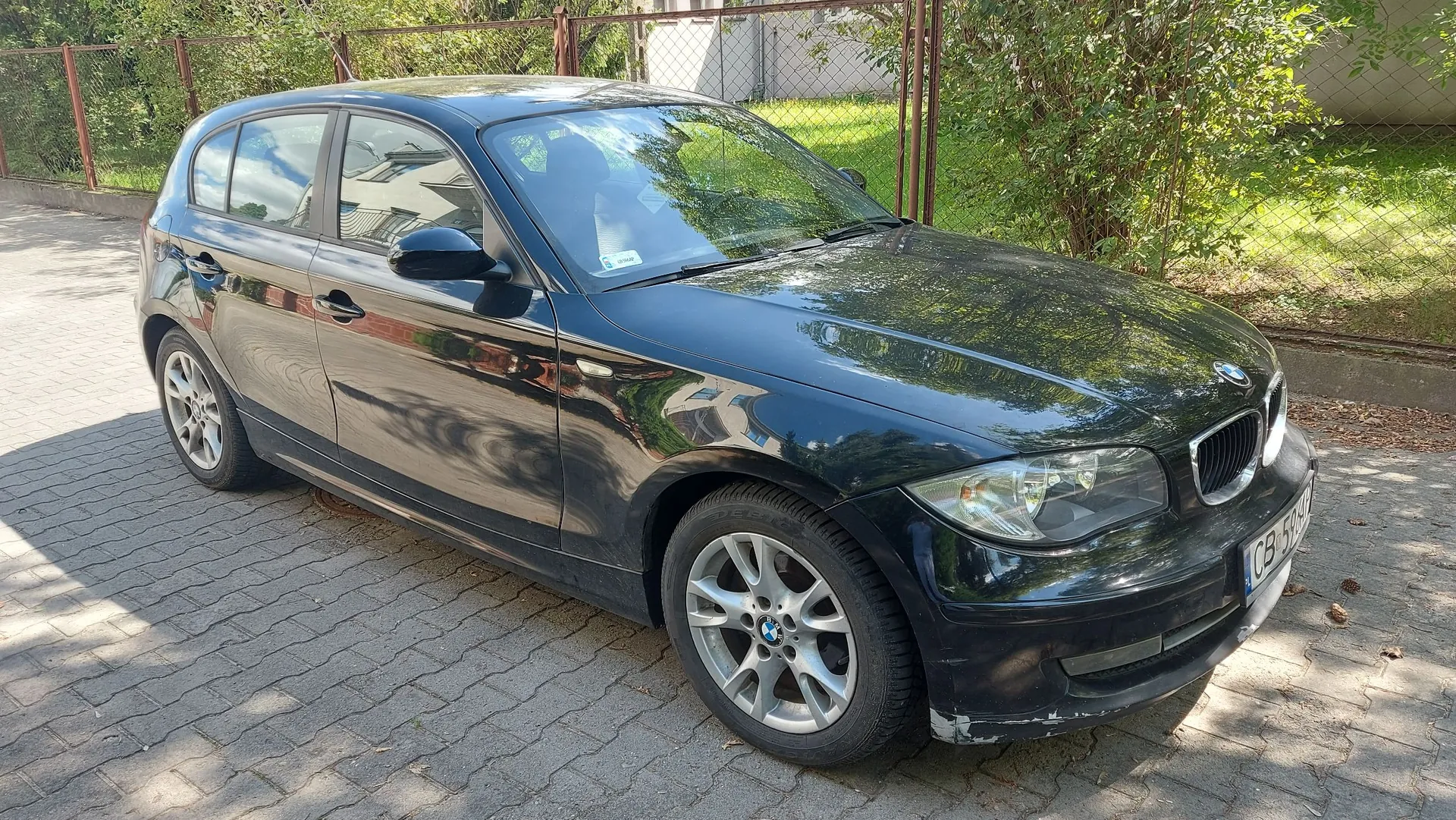 Czarny samochód marki BMW w nadwoziu hatchback, zaparkowany na kostce brukowej. Pojazd jest widoczny z przodu i z boku. W tle znajduje się metalowy płot oraz zielone krzewy i trawnik. Na przednim zderzaku widoczne są zarysowania, a na masce odbijają się drzewa.