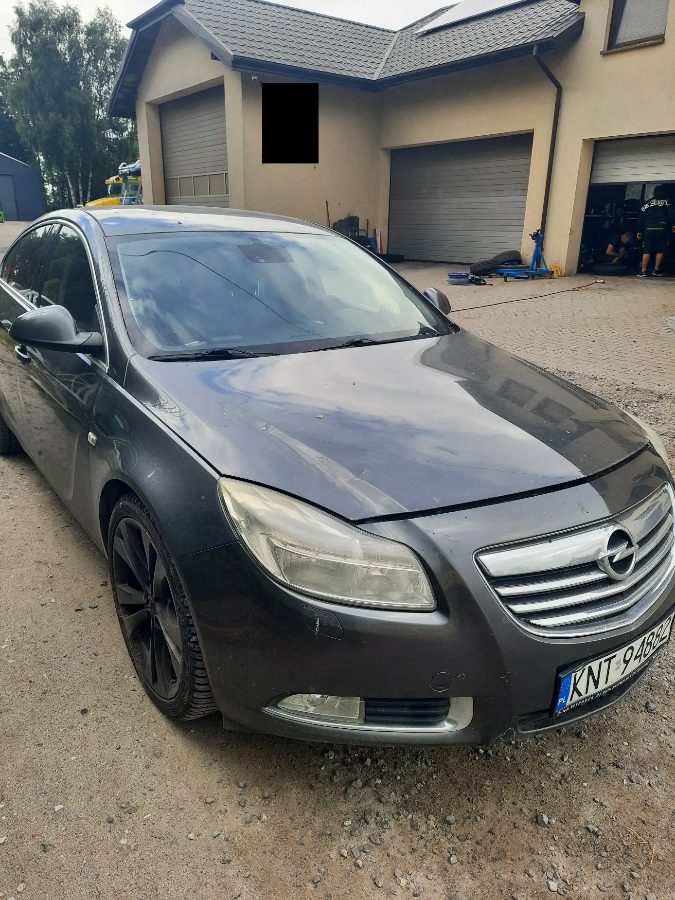 Ciemnoszary samochód marki Opel zaparkowany na piaszczystym podjeździe przed domem z dwoma garażami. Na samochodzie widoczna jest tablica rejestracyjna o numerze KNT 94882. W tle znajduje się budynek z beżową elewacją i ciemnym dachem.