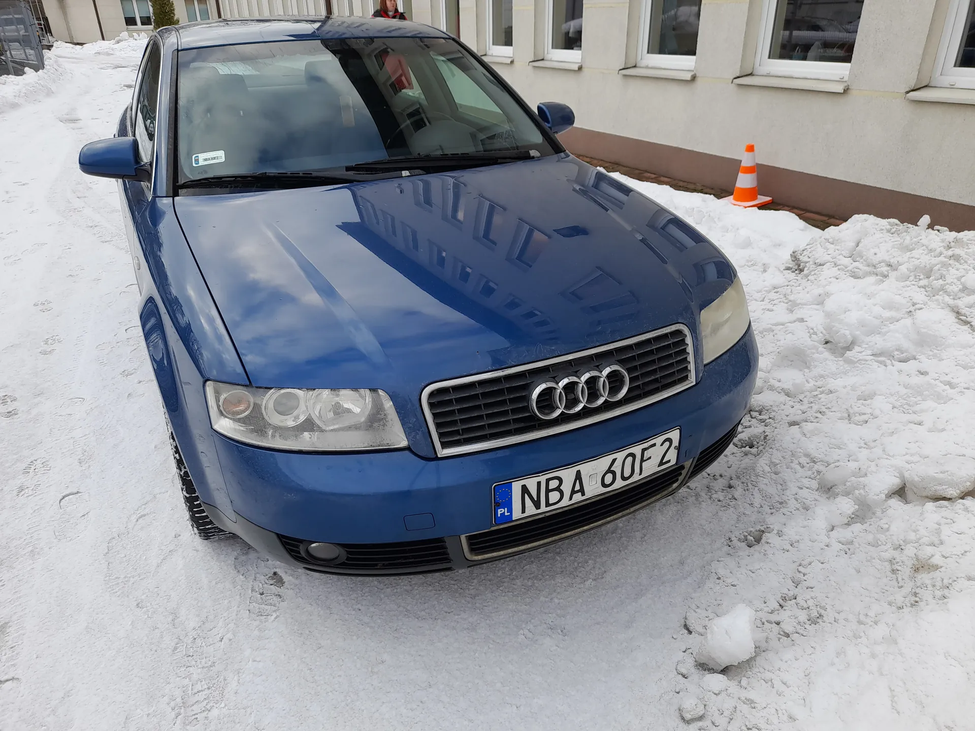 Niebieski samochód Audi z tablicą rejestracyjną NBA 60F2 (PL) zaparkowany na śnieżnej nawierzchni. W tle budynek z oknami, pomarańczowo-biały stożek drogowy oraz ośnieżone tory. Widoczne reflektory, atrapa chłodnicy z logo Audi i odbicia na karoserii.