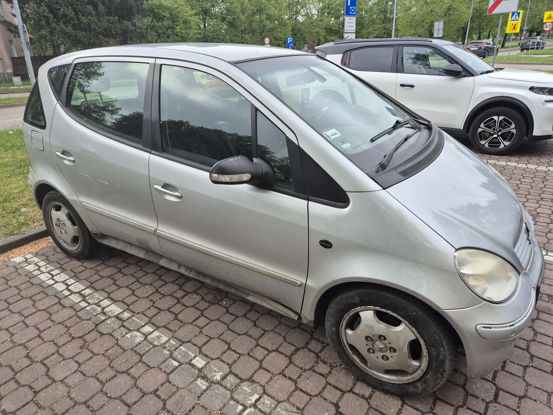 Srebrny samochód osobowy typu hatchback zaparkowany na brukowanej powierzchni. Po lewej stronie widoczna trawa i fragment budynku, w tle drzewa oraz biały SUV. Na przedniej szybie samochodu naklejki, a na masce emblemat marki. W tle znaki drogowe: niebieski znak parkingu, żółty znak ostrzegawczy i tablica z ograniczeniem prędkości 40. Samochód ma czarne felgi i widoczny napis 'Elegance' na boku.