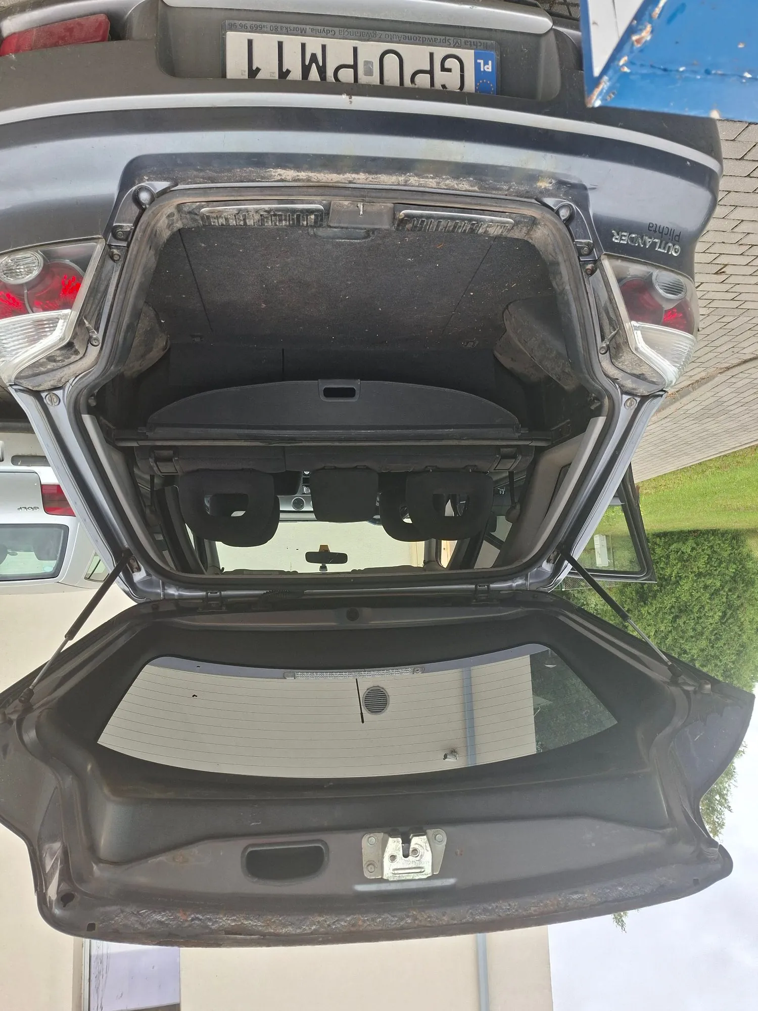 Zdjęcie otwartego bagażnika szarego samochodu marki Mitsubishi Outlander. Wewnątrz bagażnika widoczne jest ciemne obicie oraz złożone tylne siedzenia. Po prawej stronie znajduje się tablica rejestracyjna o numerze GPU PM11 z oznaczeniem PL. Obraz jest zorientowany poziomo, mimo że przedstawia tył samochodu.