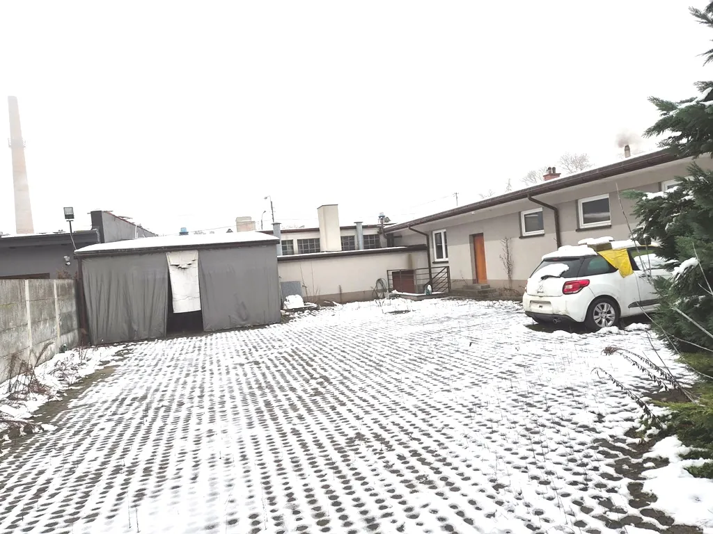 Zimowy widok na podwórze pokryte śniegiem. Na pierwszym planie znajduje się parking z betonowych kratownic wypełnionych śniegiem. Po lewej stronie stoi szary, tymczasowy budynek lub garaż z materiału płóciennego. W tle widoczny jest parterowy, jasny budynek mieszkalny z brązowymi drzwiami i oknami. Po prawej stronie zaparkowany jest biały samochód typu SUV, częściowo przykryty śniegiem. W oddali, po lewej stronie, widoczny jest wysoki, betonowy komin przemysłowy.