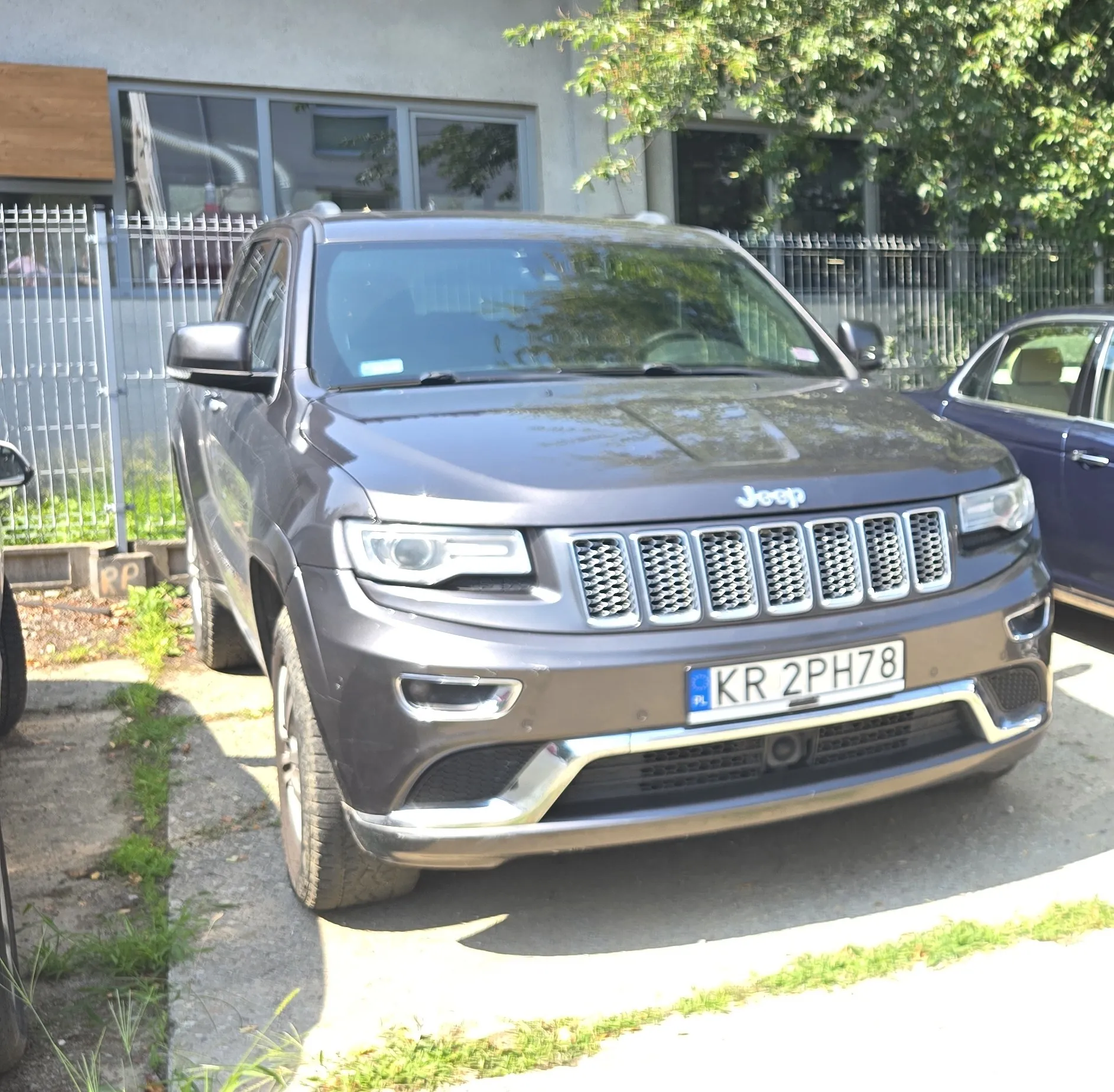 Srebrno-szary samochód marki Jeep Grand Cherokee zaparkowany na betonowym podjeździe przed budynkiem z dużymi oknami i metalowym ogrodzeniem. Pojazd ma polską tablicę rejestracyjną o numerze KR 2PH78.