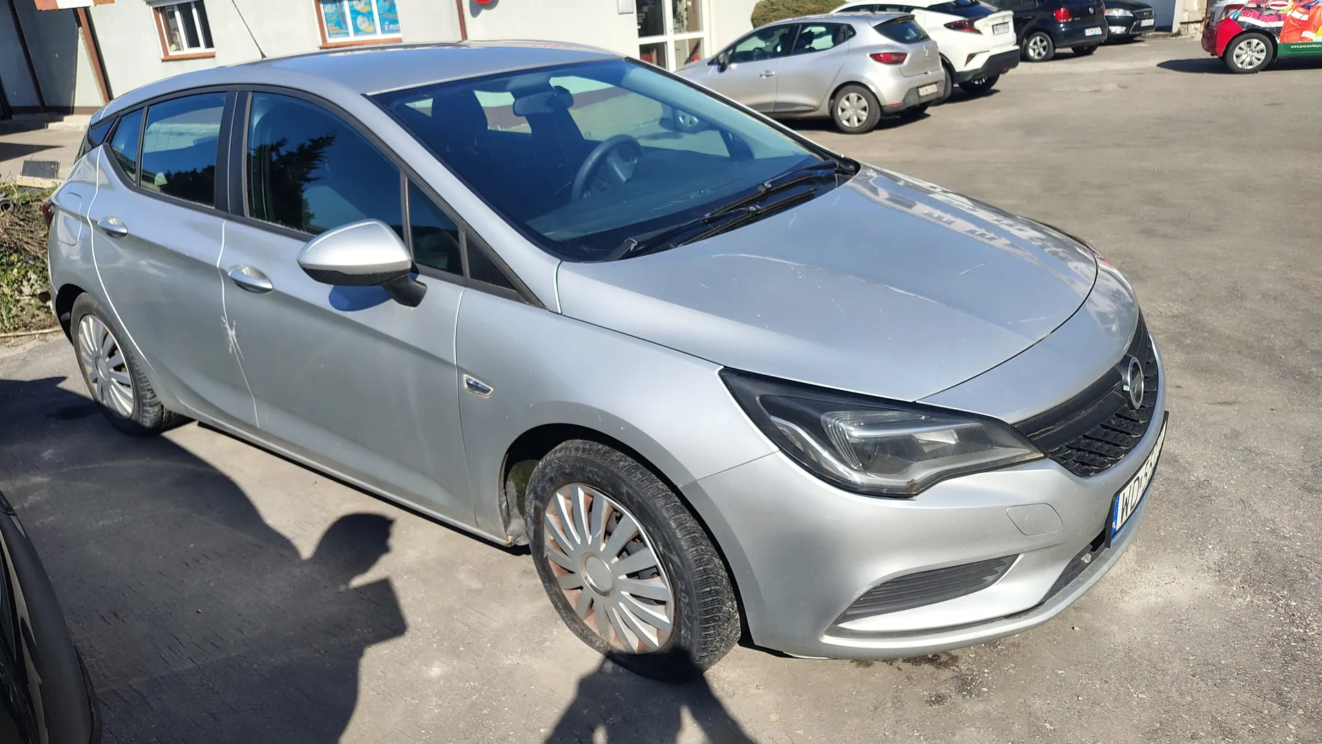 Srebrny samochód osobowy typu hatchback zaparkowany na asfaltowym placu parkingowym. W tle widoczne są inne pojazdy, budynek z oknami i drzwiami wejściowymi oraz fragmenty tablic reklamowych. Na pierwszym planie cień osoby robiącej zdjęcie. Samochód ma czystą karoserię, felgi aluminiowe oraz tablicę rejestracyjną z nieczytelnym numerem. Oświetlenie słoneczne rzuca cienie na powierzchnię parkingu.