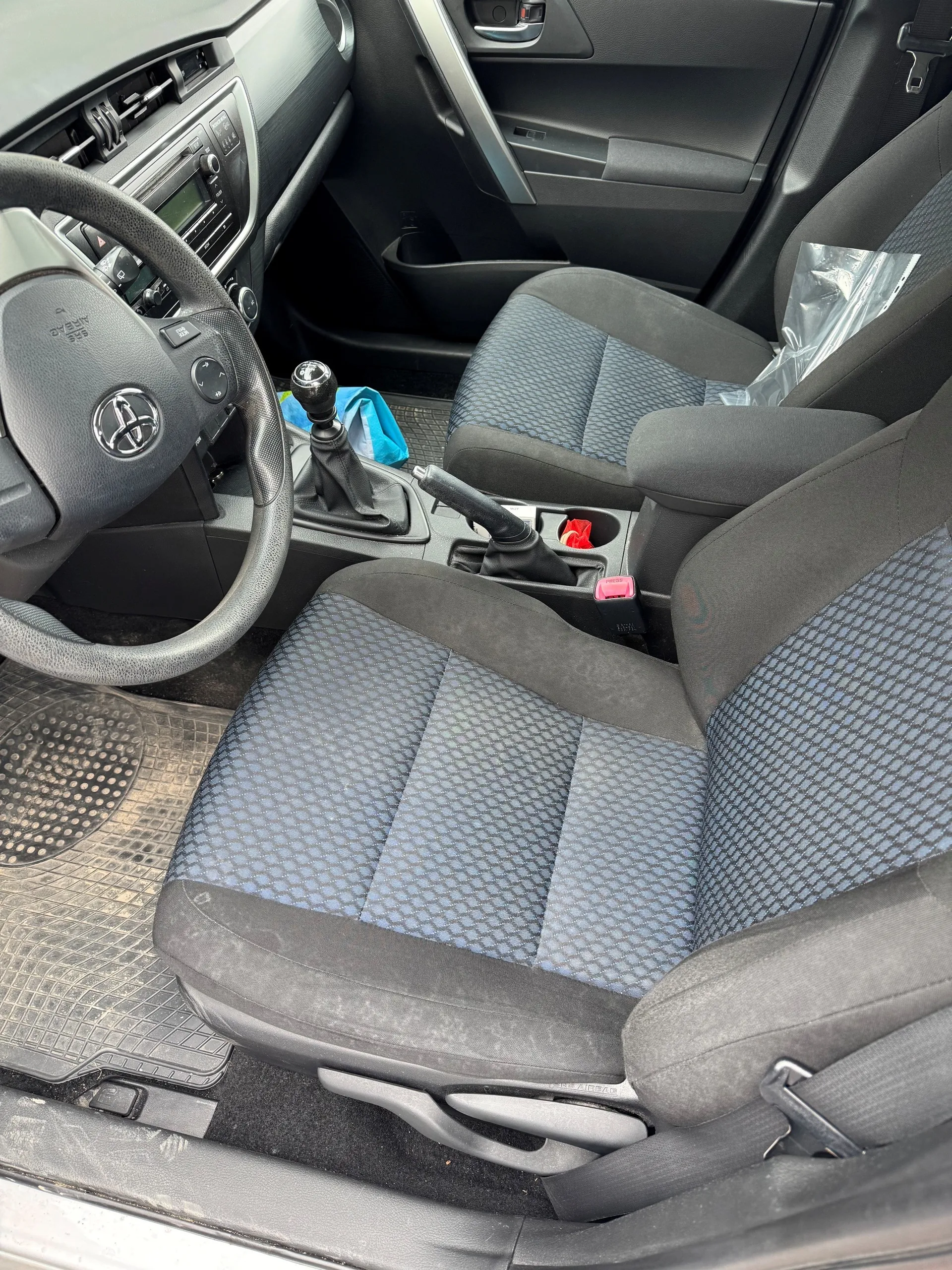 Wnętrze samochodu marki Toyota z widocznym kierownicą z logo, deska rozdzielcza z ekranem i przyciskami, dwa przednie fotele z niebiesko-szarym wzorem, dźwignia zmiany biegów, hamulec ręczny, niebieska torba na podłodze oraz czerwony przedmiot w uchwycie na napoje. Na fotelu pasażera leży przezroczysta folia. Podłoga wyłożona jest gumowymi dywanikami.