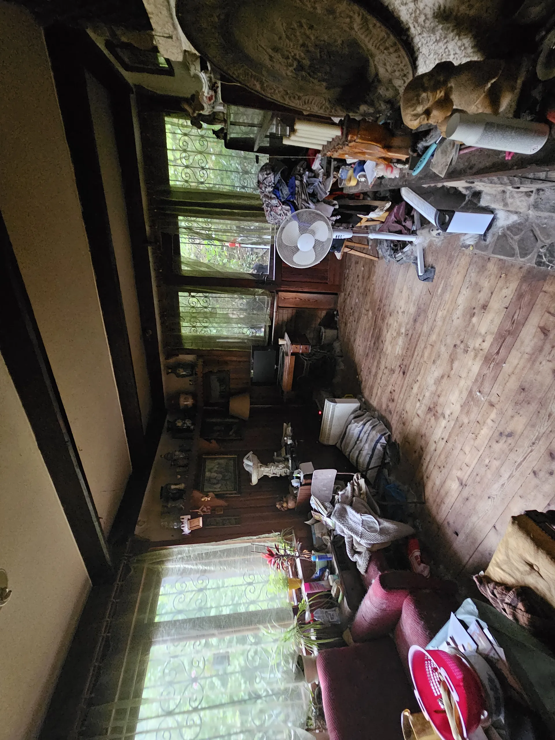 Zdjęcie przedstawia przestronny, zatłoczony pokój z drewnianym sufitem i kamienną ścianą. W pomieszczeniu znajdują się trzy okna z żelaznymi kratami i zasłonami, biały wentylator sufitowy, telewizor, regały z różnymi przedmiotami oraz platforma lub łóżko w centralnej części. Na ścianach wiszą dekoracje, a na podłodze i powierzchniach leżą różne przedmioty, w tym worki, ubrania i naczynia. Przez okna widać zielony teren na zewnątrz.