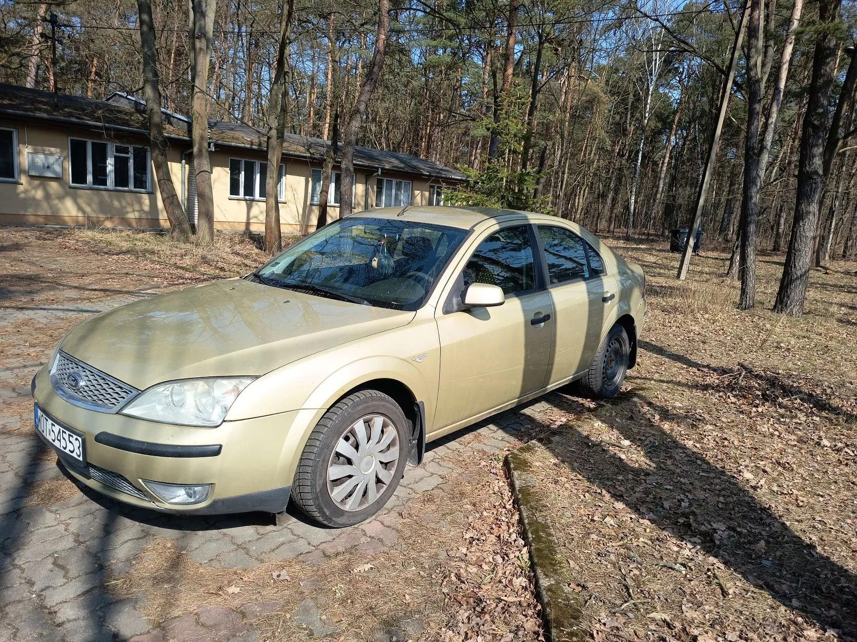 Złoty samochód osobowy marki Ford, model Mondeo, zaparkowany na brukowanej powierzchni. Widoczna tablica rejestracyjna KOT 54553. W tle jednopiętrowy budynek o beżowej elewacji z kilkoma oknami, otoczony wysokimi drzewami bez liści. Podłoże pokryte suchymi liśćmi, słoneczny dzień z wyraźnymi cieniami. Samochód stoi pod kątem do obserwatora, z przodu skierowanym w lewą stronę kadru.
