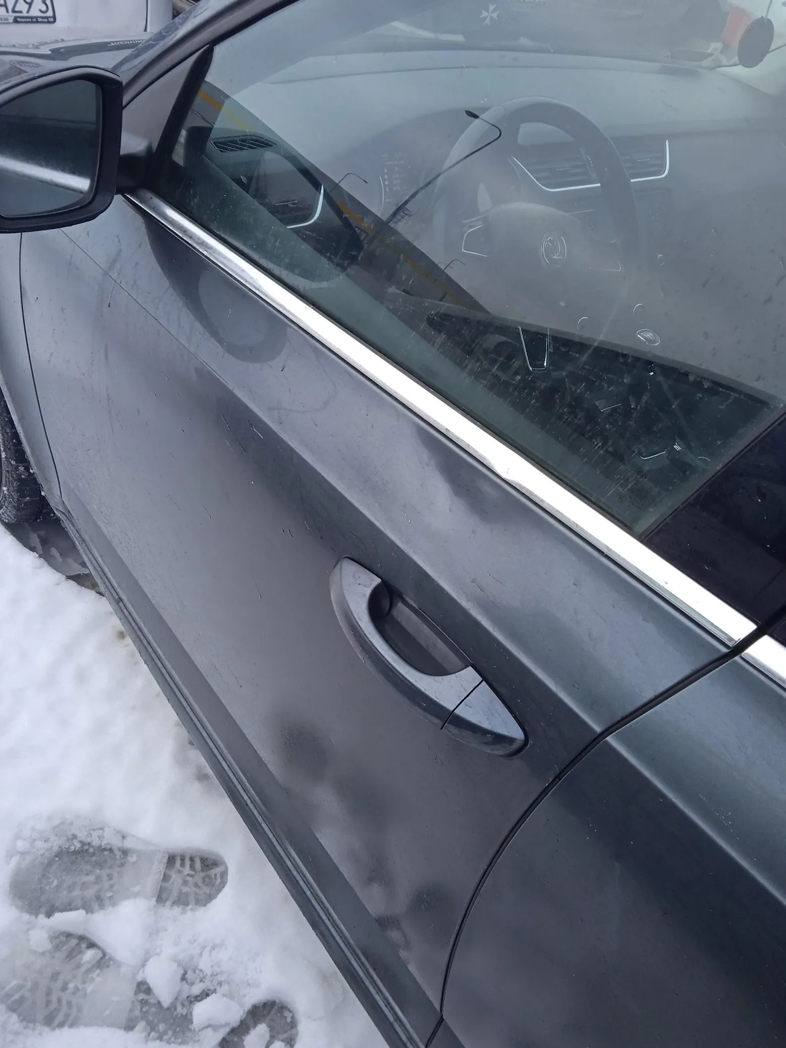 Zbliżenie na bok ciemnoszarego samochodu marki Skoda, zaparkowanego na śniegu. Widoczne są drzwi kierowcy z klamką, lusterko boczne oraz wnętrze auta przez szybę, w tym kierownica z logo marki. Na śniegu obok samochodu widoczne są odciski butów.