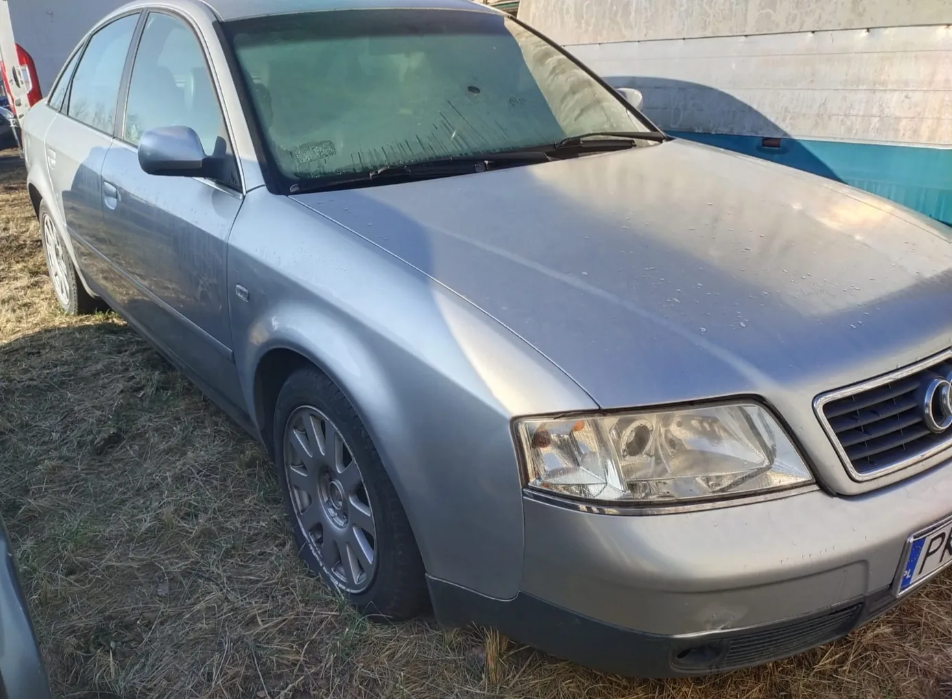 Srebrny samochód osobowy marki Audi, czterodrzwiowy sedan, zaparkowany na suchym trawniku. Widoczna część tablicy rejestracyjnej z literami PK. W tle betonowa ściana z niebieskim pasem. Samochód ma srebrne felgi, przednie światła oraz zarys logo na atrapie chłodnicy.