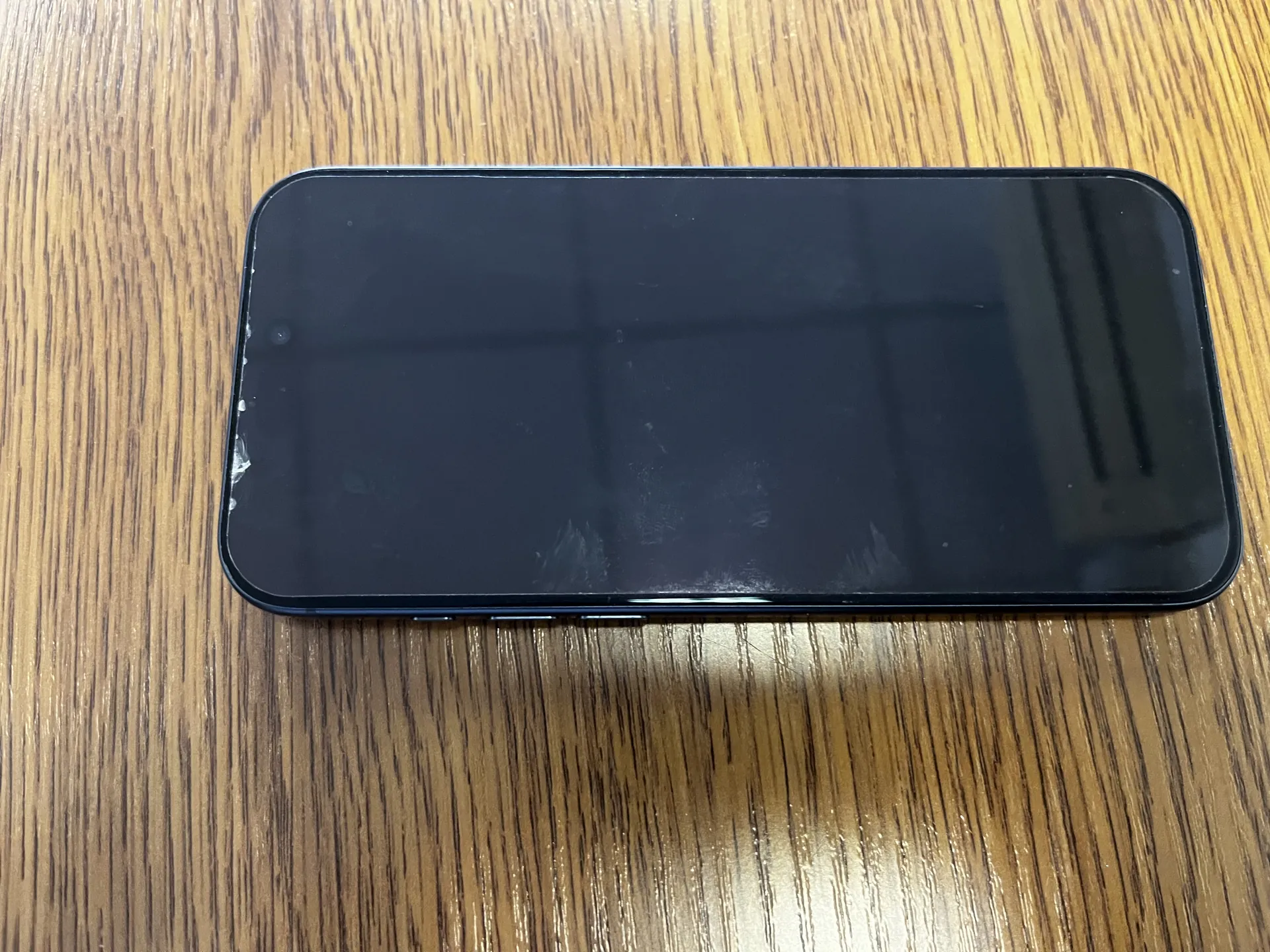 Czarny smartfon z wyłączonym ekranem leży poziomo na drewnianym stole o widocznych żyłkach. Ekran posiada ślady palców i odbicia światła. W lewym górnym rogu ekranu znajduje się mały otwór pod kamerę. Stół ma jasnobrązowy kolor z ciemnymi żyłkami.