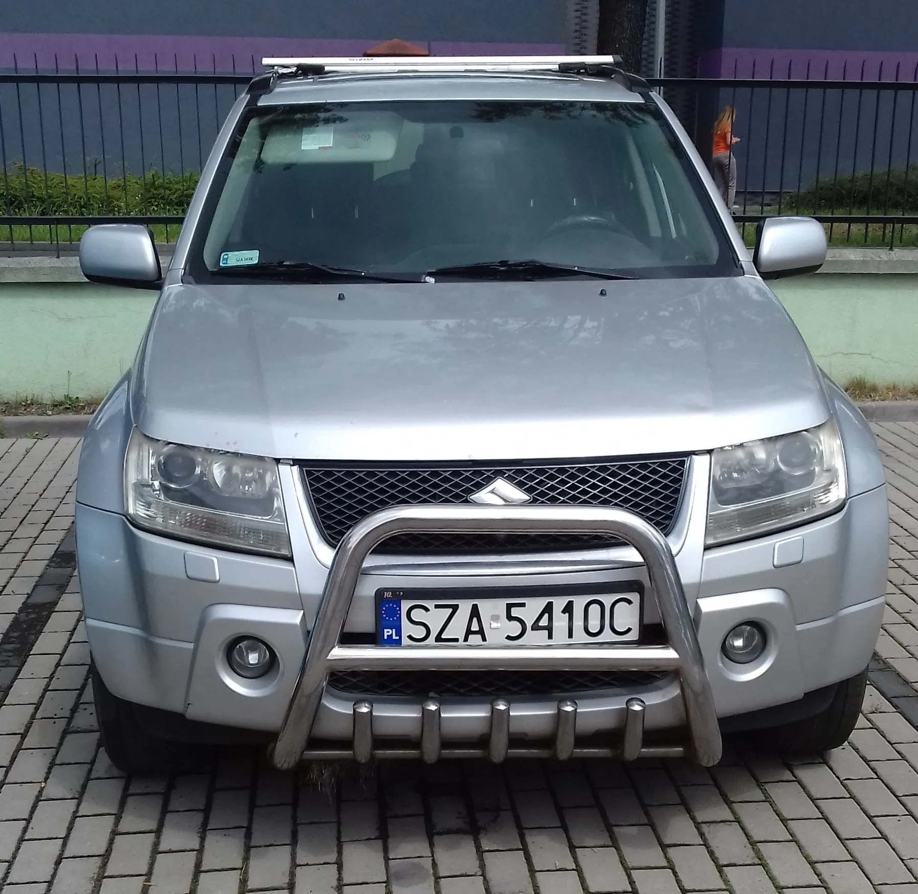 Srebrny samochód marki Mitsubishi widziany od przodu, zaparkowany na kostce brukowej. Pojazd posiada zamontowany na przednim zderzaku metalowy ochraniacz (bullbar) oraz bagażnik dachowy. Tablica rejestracyjna samochodu to SZA 5410C.
