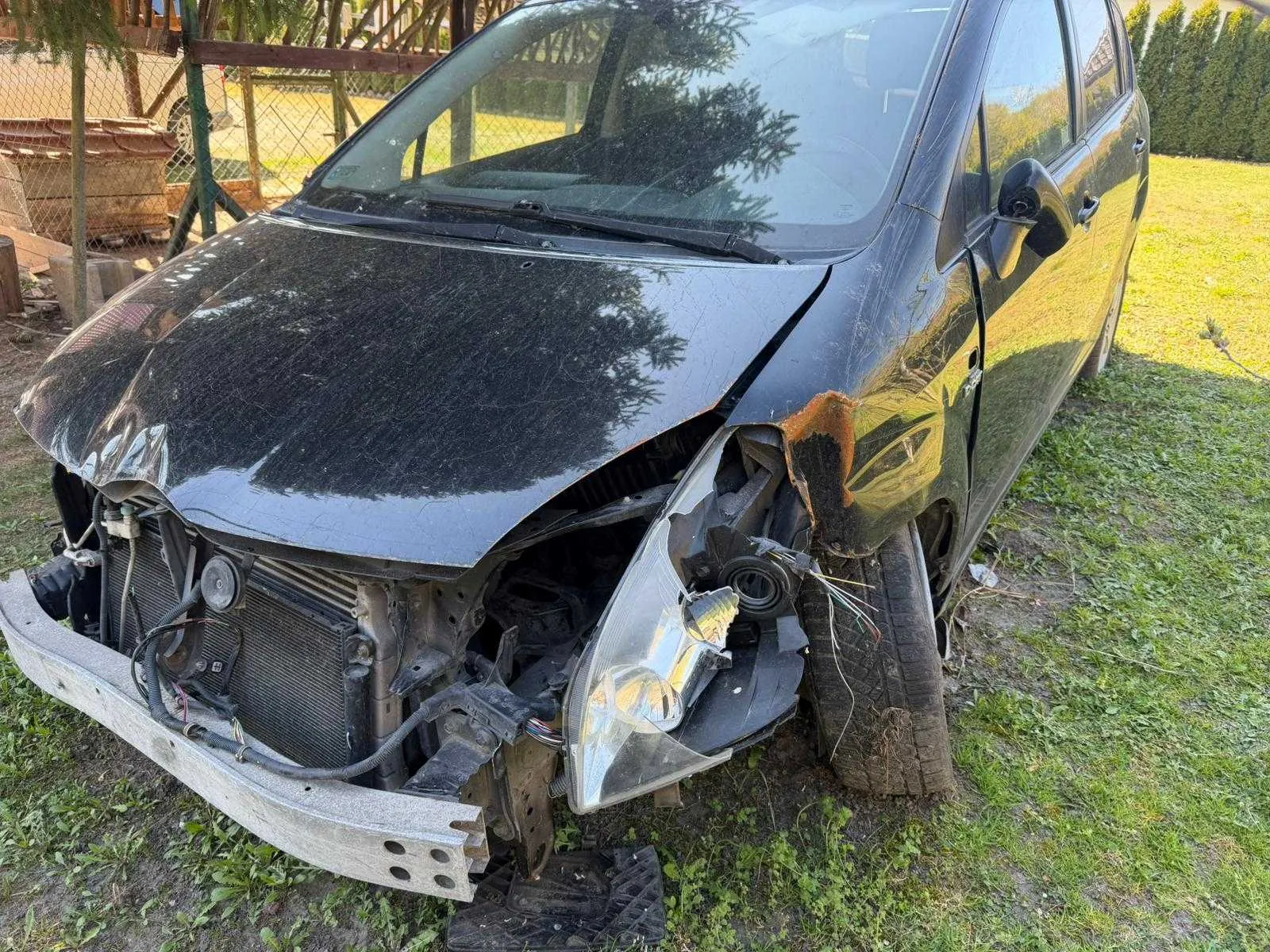Zniszczony czarny samochód osobowy z uszkodzonym przodem. Widoczne są pęknięte elementy karoserii, zdeformowana maska, uszkodzony zderzak, odsłonięte części silnika oraz pęknięte światło przednie. Samochód stoi na trawie, w tle ogrodzenie siatkowe, drewniane konstrukcje i krzewy. Oświetlenie wskazuje na dzień.