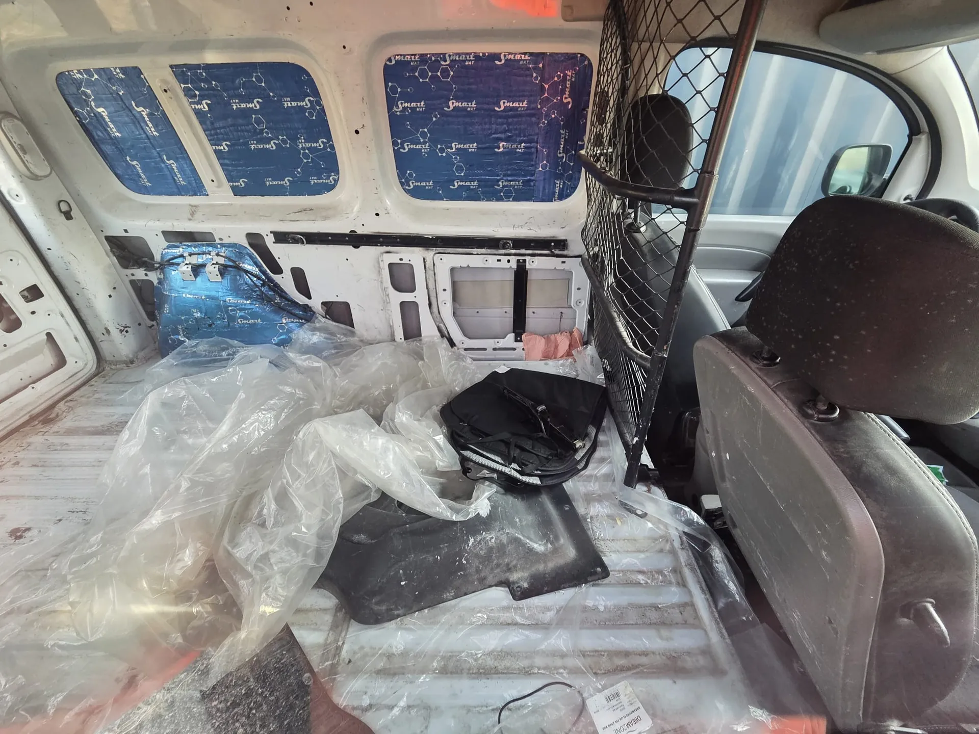 Wnętrze furgonetki z widocznymi elementami: białe ściany z niebieskimi panelami okiennymi pokrytymi powtarzającym się napisem "Smart" oraz wzorem cząsteczek chemicznych, metalową siatką oddzielającą przestrzeń pasażerską od ładunkowej, zniszczonymi siedzeniami kierowcy i pasażera, przezroczystymi foliami na podłodze oraz czarną torbą i innymi przedmiotami na ładowni. W tle widoczne są okna z niebieskimi panelami i fragment zewnętrzny za szybą.