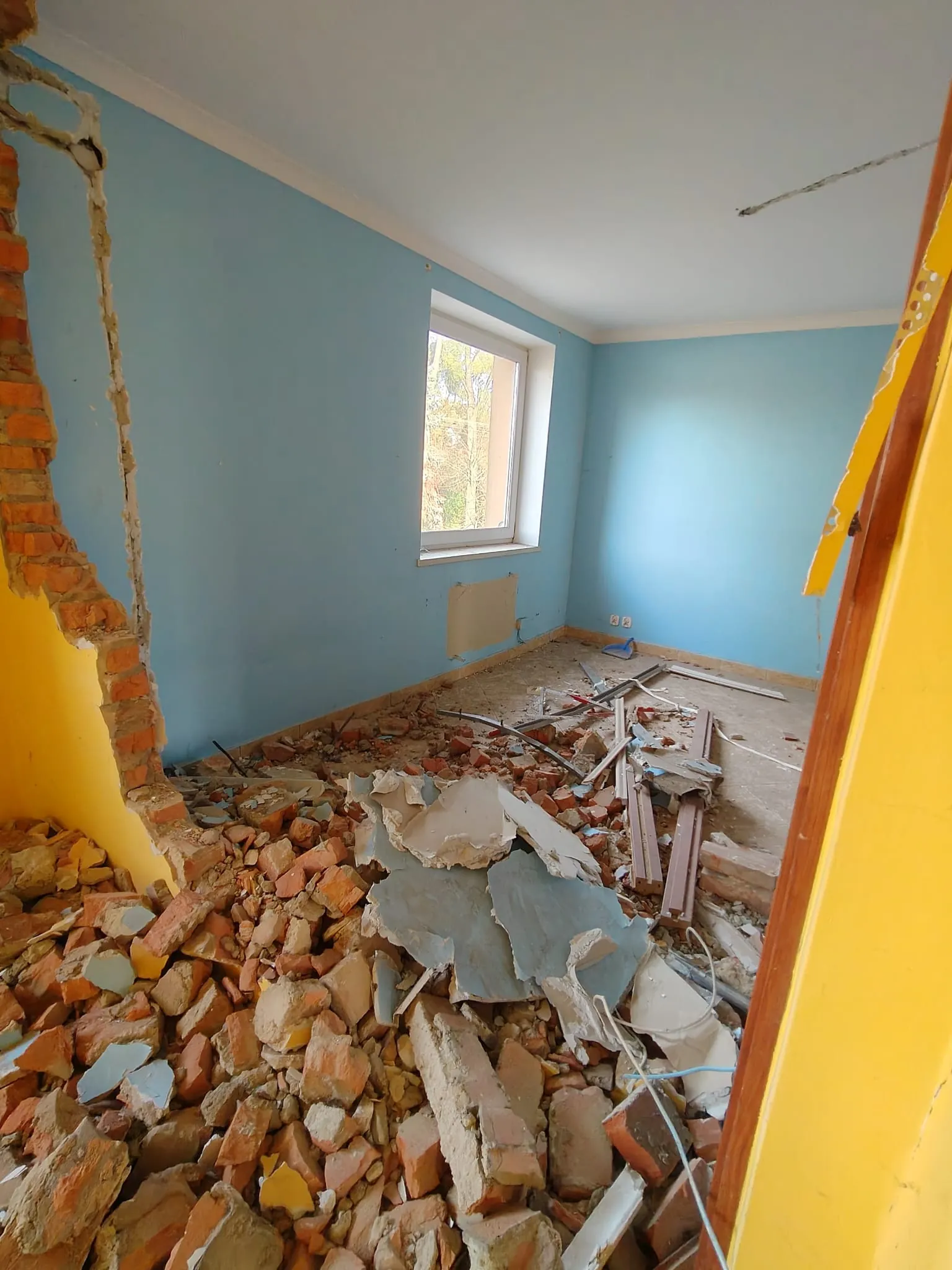 Wnętrze pomieszczenia w trakcie remontu. Ściany są pomalowane na jasnoniebieski kolor, a jedna z nich jest żółta. W centrum znajduje się okno. Na podłodze leży duża ilość gruzu, consisting z czerwonych cegieł, kawałków tynku i betonowych odłamków. Po lewej stronie widoczny jest wyłom w ścianie odsłaniający ceglaną konstrukcję.