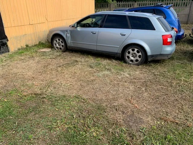 Srebrny samochód typu kombi zaparkowany bokiem na trawiastym terenie z wysuszoną trawą. Samochód ma widoczne uszkodzenie nad przednim lewym kołem. W tle znajduje się żółta ściana budynku oraz fragment niebieskiego samochodu zaparkowanego za srebrnym autem.
