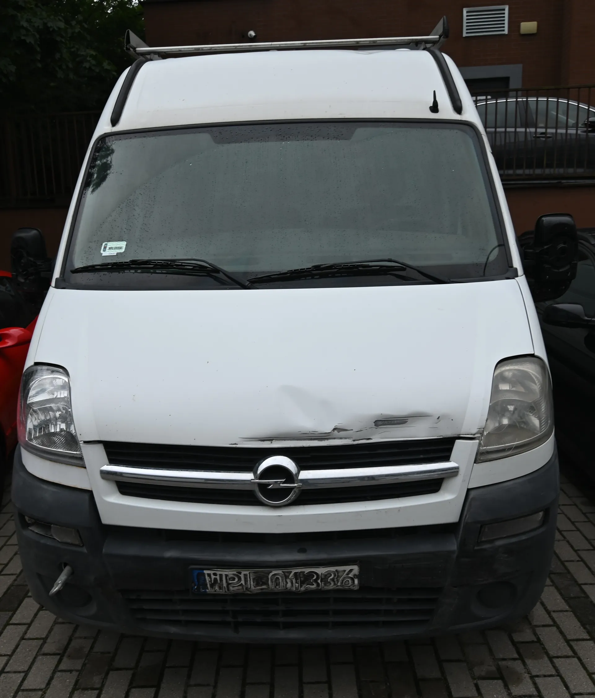 Widok z przodu białego vana marki Opel. Pojazd ma wgniecenie na masce silnika po prawej stronie (patrząc od przodu). Na przednim zderzaku znajduje się tablica rejestracyjna o numerze HPL 01386. Na dachu zamontowany jest srebrny bagażnik. Samochód stoi na kostce brukowej.