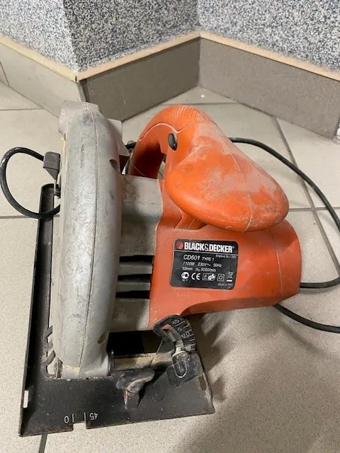 Czerwono-szara piła tarczowa marki Black & Decker model CD061 o mocy 1100W, napięciu 230V i częstotliwości 50Hz. Urządzenie leży na podłodze z białymi płytkami, w tle widoczna jest szara, kropkowana ściana. Na piłce znajduje się czarny kabel zasilający oraz czarna podstawa z oznaczeniami kątowymi (0, 45).