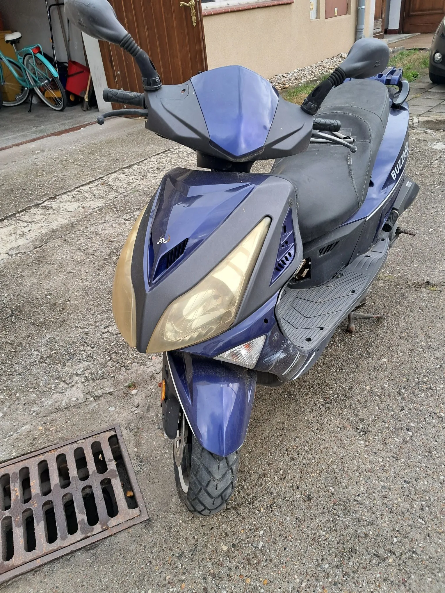 Niebieski skuter zaparkowany na betonowym podłożu. Widoczny jest przód pojazdu z dużym reflektorem, kierownica z lusterkami oraz czarne siedzenie. W tle znajduje się fragment budynku i metalowa kratka ściekowa.