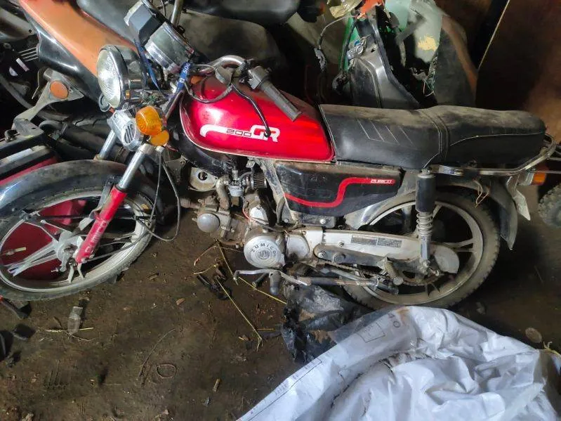 Czerwony motocykl marki Suzuki z napisem "200 CR" na zbiorniku paliwa, czarnym siedzeniem i widocznymi częściami silnika. Motocykl znajduje się w pomieszczeniu z brudną podłogą, otoczony innymi pojazdami i częściami mechanicznymi. W tle widoczne są fragmenty innych motocykli oraz biała folia na ziemi.