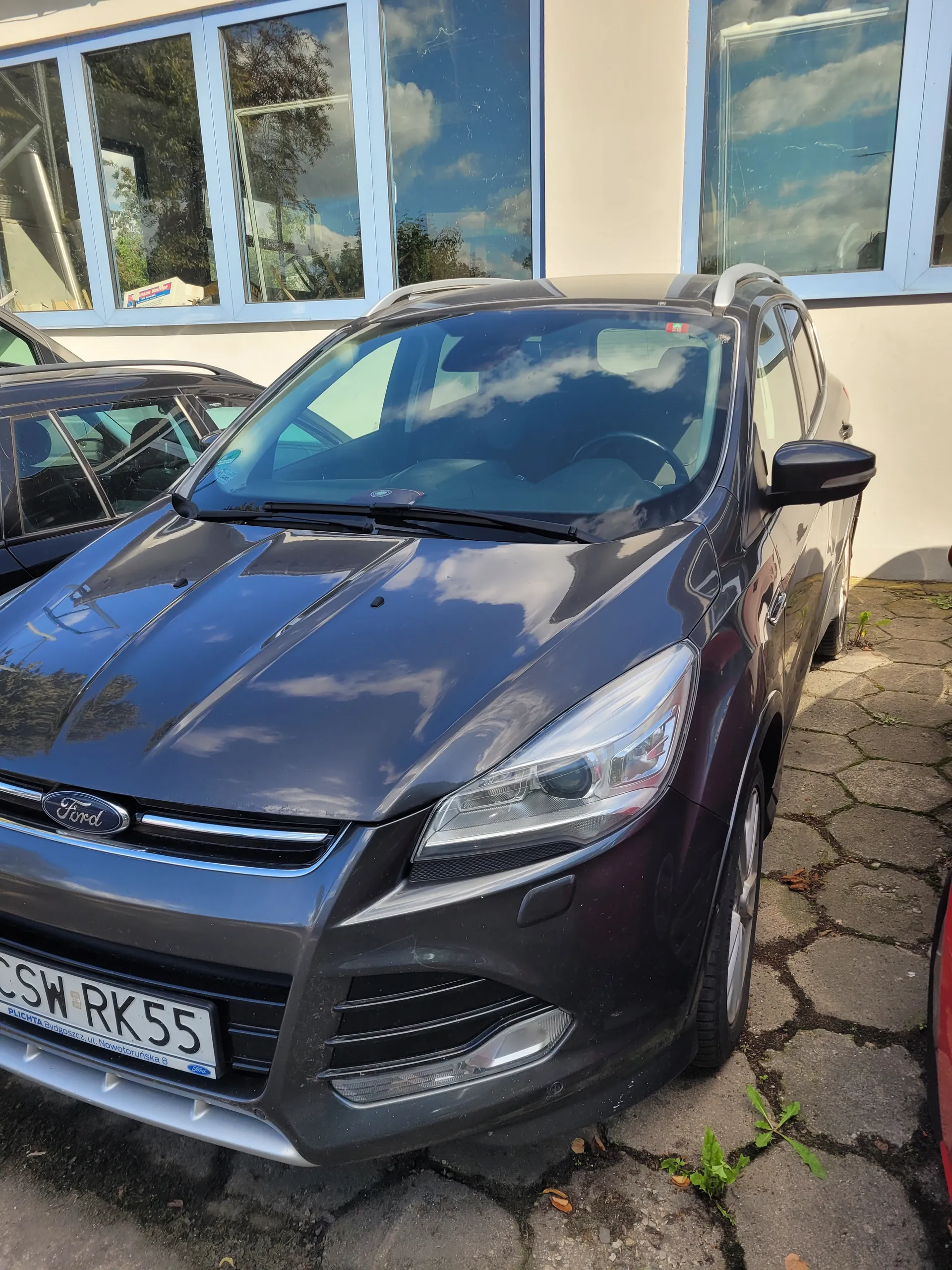 Ciemnoniebieski samochód marki Ford zaparkowany na brukowanej powierzchni. Tablica rejestracyjna z napisem SW RK55 umieszczona pionowo. W tle budynek z niebieskimi ramami okiennymi odbijający niebo i drzewa. Na ziemi widoczne są kamienie brukowe oraz małe rośliny.